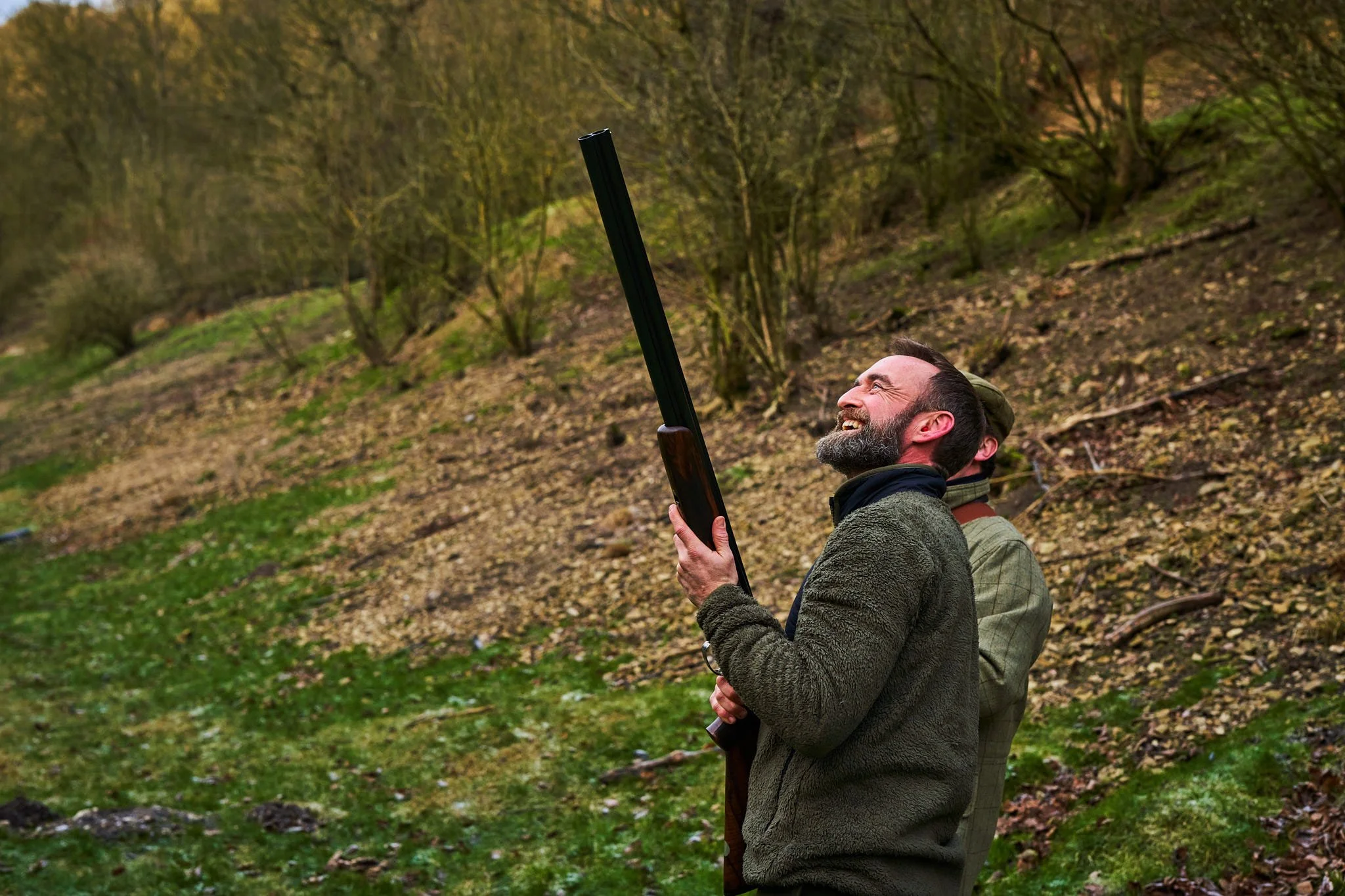 A man in outdoor clothing holding a shotgun, smiling and looking upward in a wooded area during daytime.
