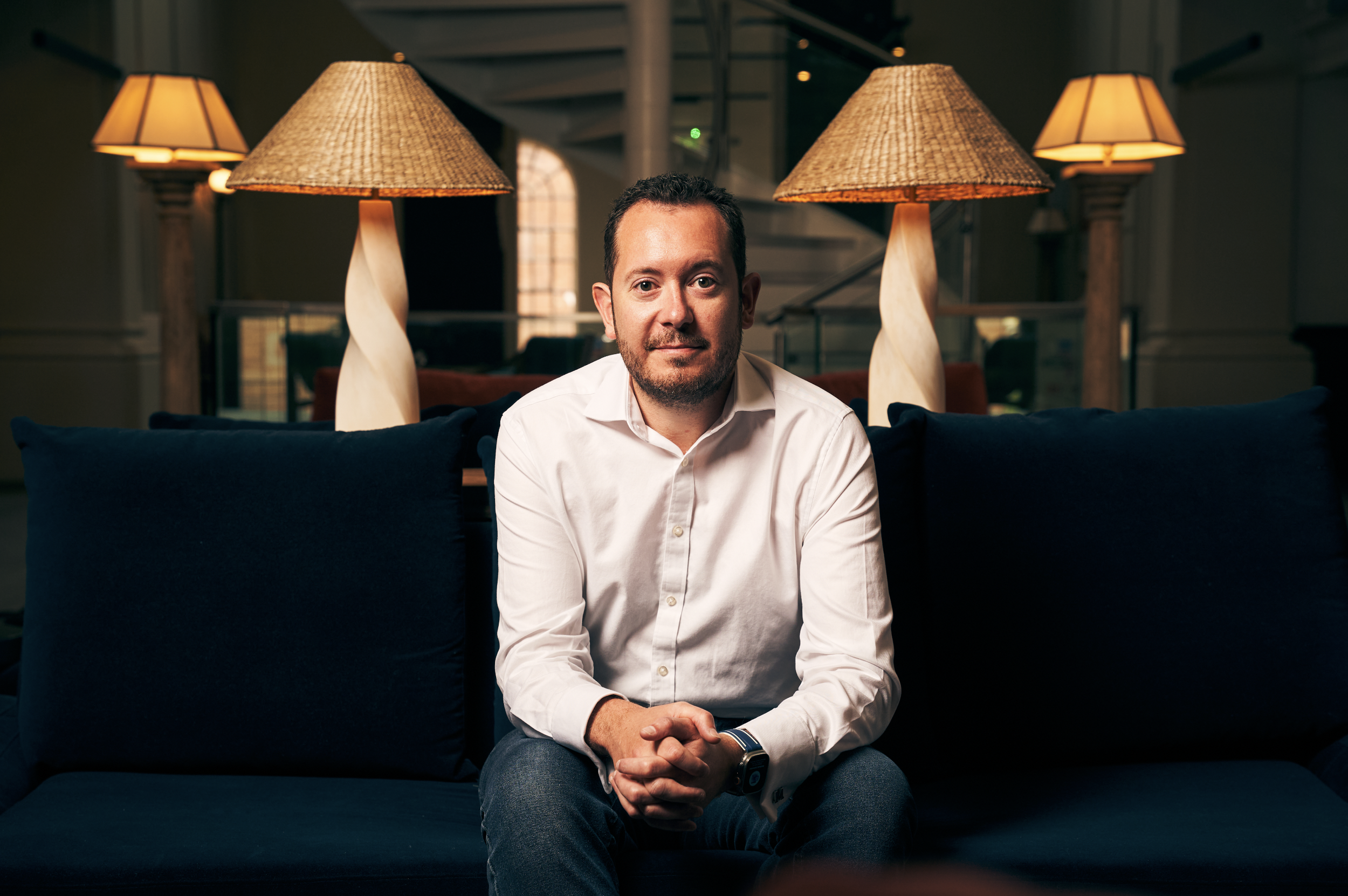 A man in a white shirt sitting on a dark blue sofa with pillows in a warmly lit room, with lamps and a staircase in the background.