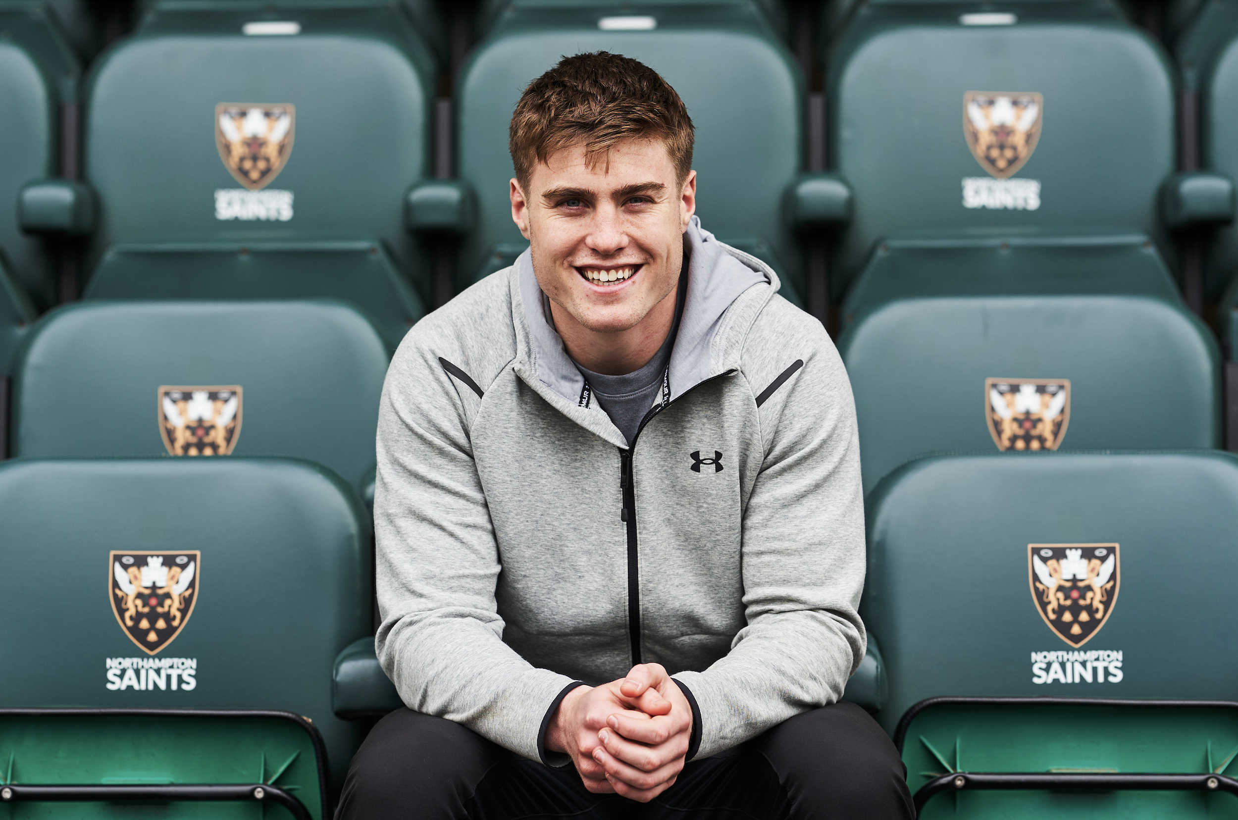 A young man is sitting on stadium seats with the Northampton Saints logo. He is smiling, wearing a gray Under Armour hoodie and black pants, with his hands clasped together.