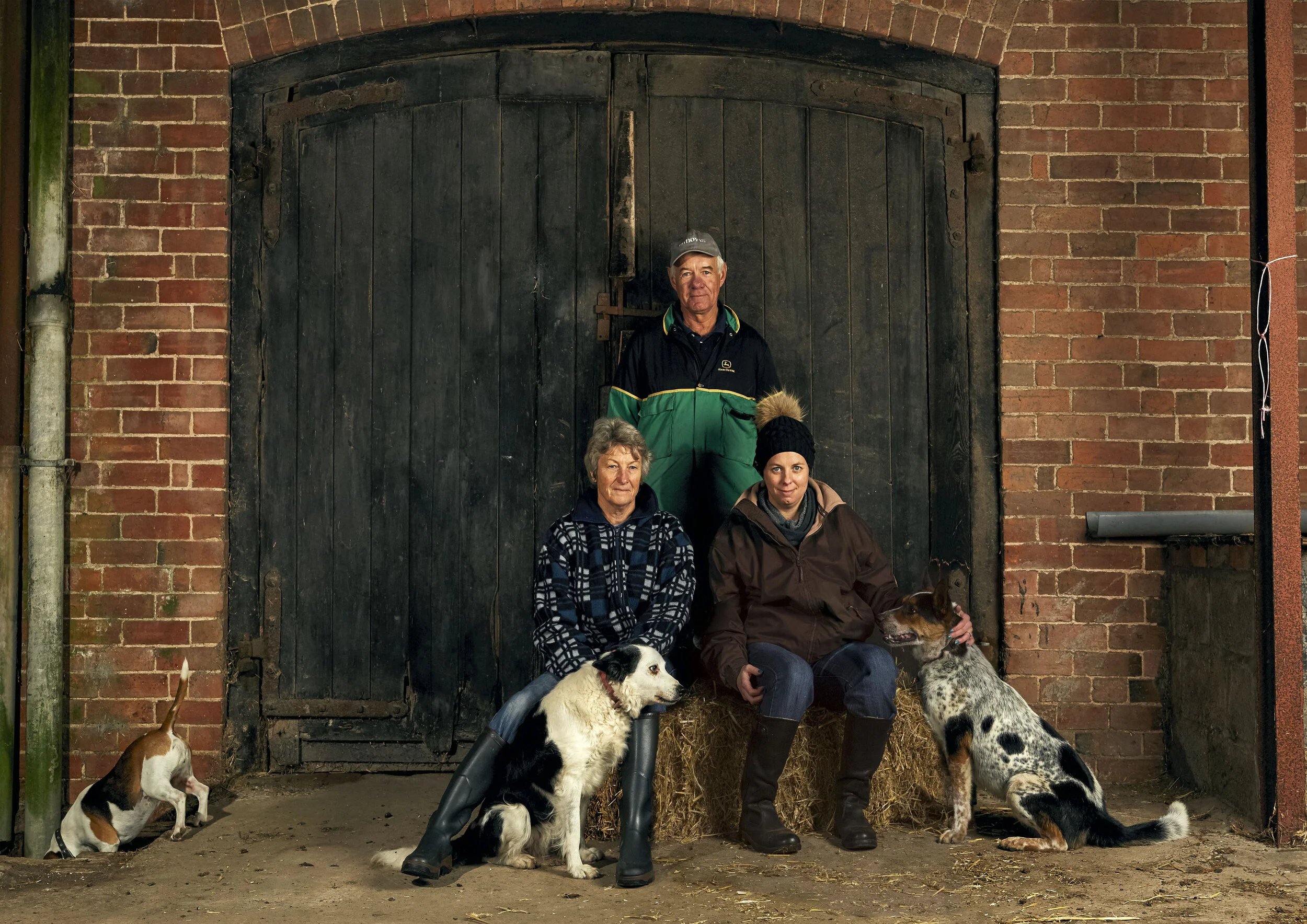 A group of four people and five dogs sitting and standing in front of a black barn door with brick walls on either side. The scene is rustic and outdoors.