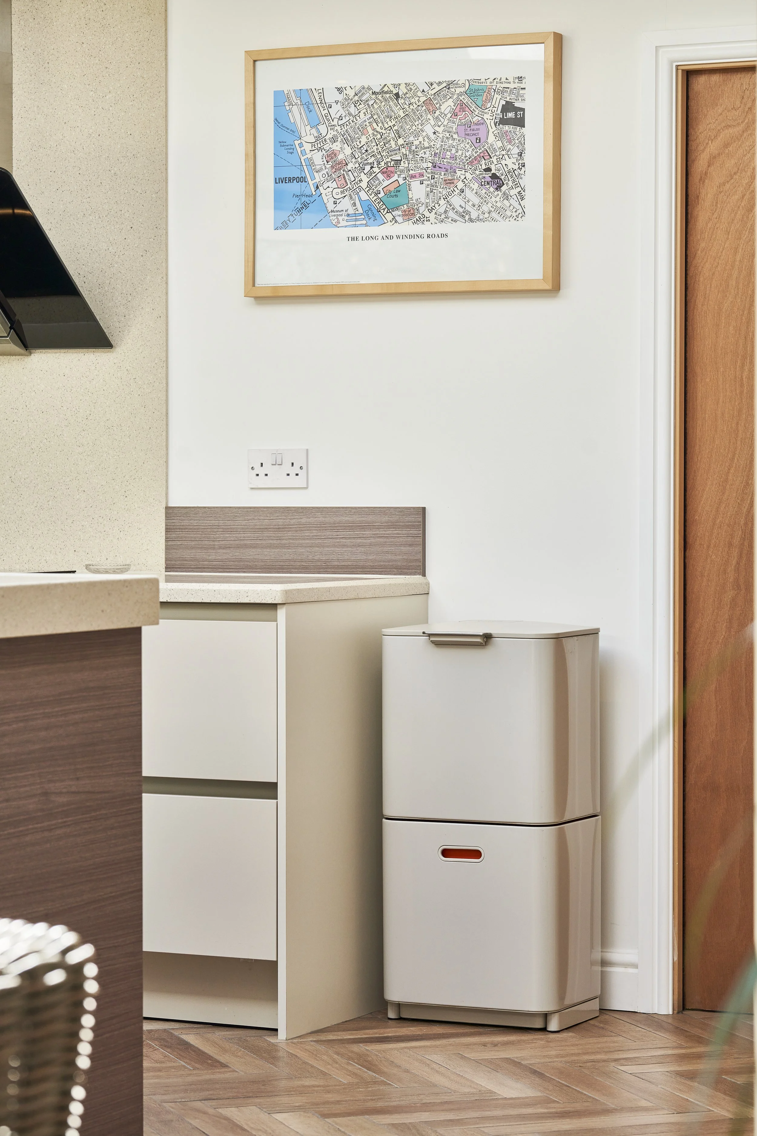 A small white refrigerator next to a cabinet in a kitchen, with a framed map of Liverpool hanging on the wall above. There is also a power outlet on the wall.