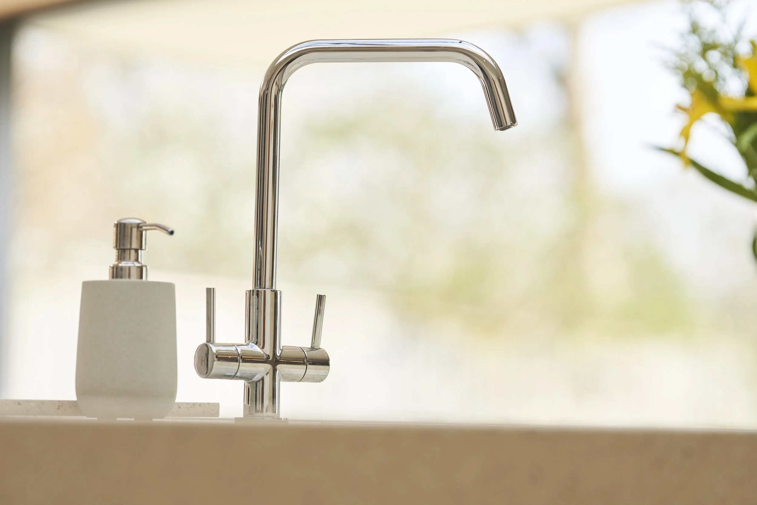 Kitchen sink with chrome faucet and a soap dispenser on a light-colored countertop, with a blurry window and greenery in the background.