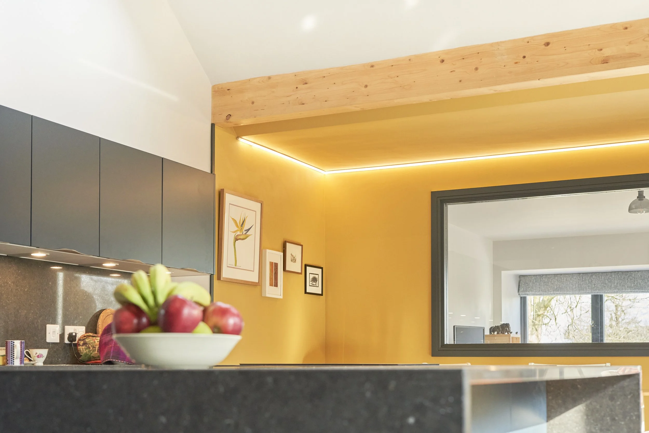 Kitchen with a bowl of assorted fruits on a dark countertop, yellow wall with framed artwork, gray cabinets, and a window reflecting a tree outside.