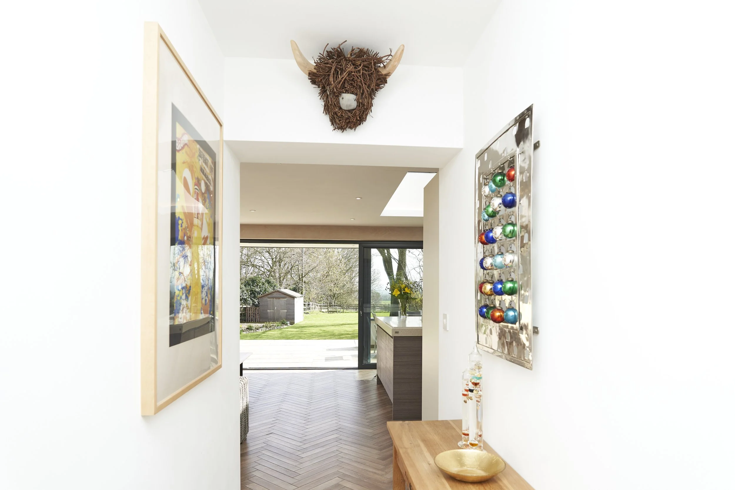 Interior hallway leading to a living space with sliding glass doors showing a garden outside, decorated with a buffalo head wall mount, colorful artwork, and a metal display with decorative balls.