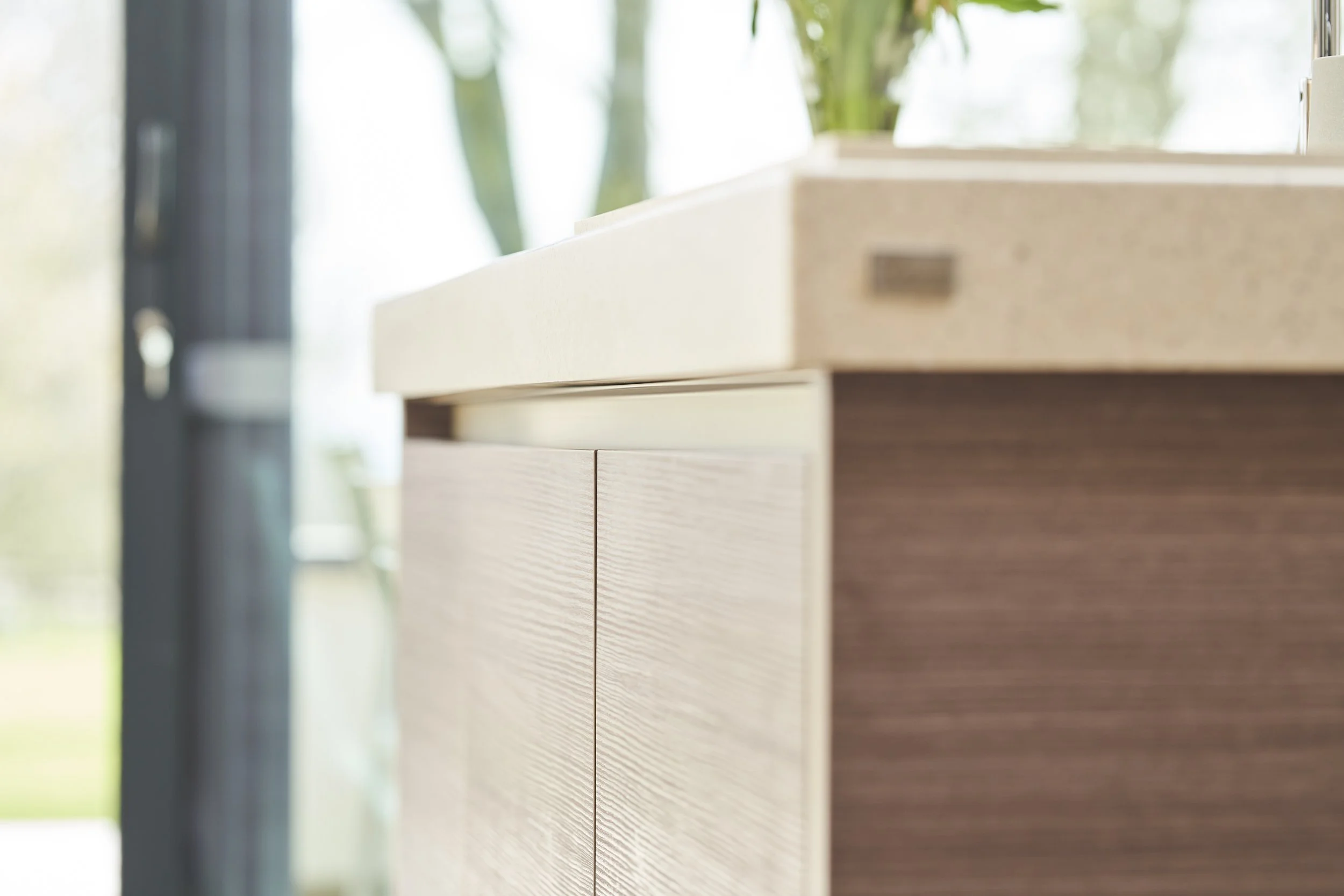 Close-up of a wooden cabinet with a beige countertop and a potted plant in the background, near a large window.