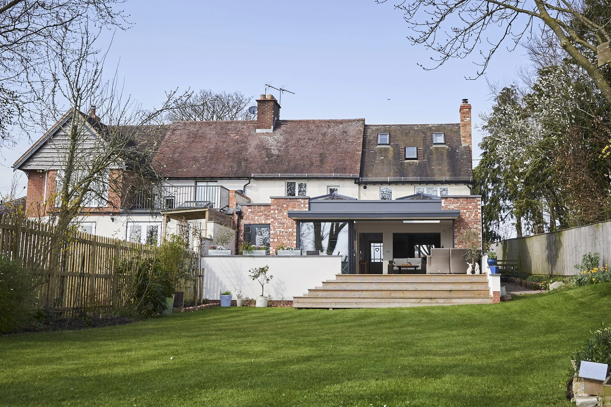 Back of a house with a large lawn, wooden deck, and multiple levels of brick and wood siding, under a clear sky.