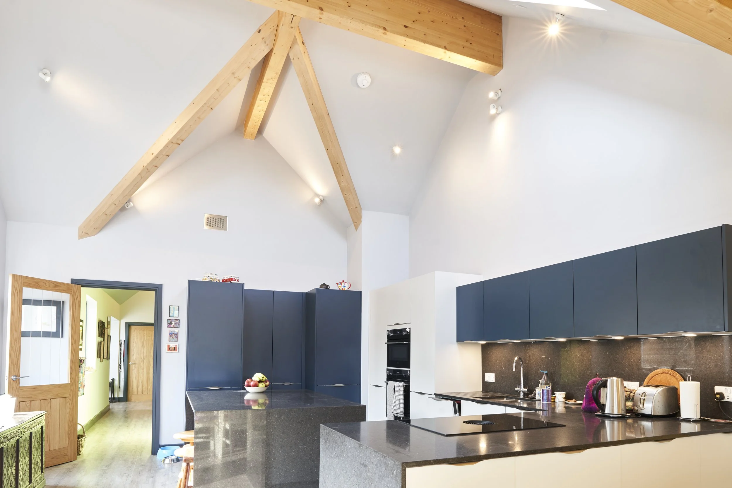 Modern kitchen with dark blue upper cabinets, black countertops, and exposed wooden beams on a high white ceiling, illuminated by spotlights.