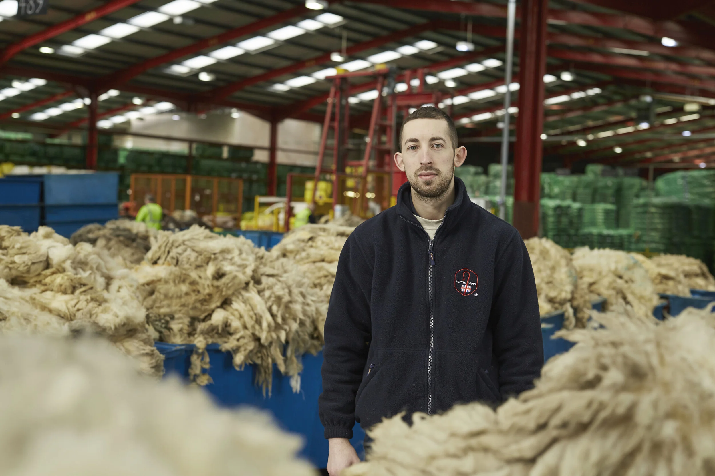A young man with short dark hair and a beard wearing a black jacket with a UK wool badge, standing in a wool processing warehouse surrounded by large piles of raw wool and industrial equipment.