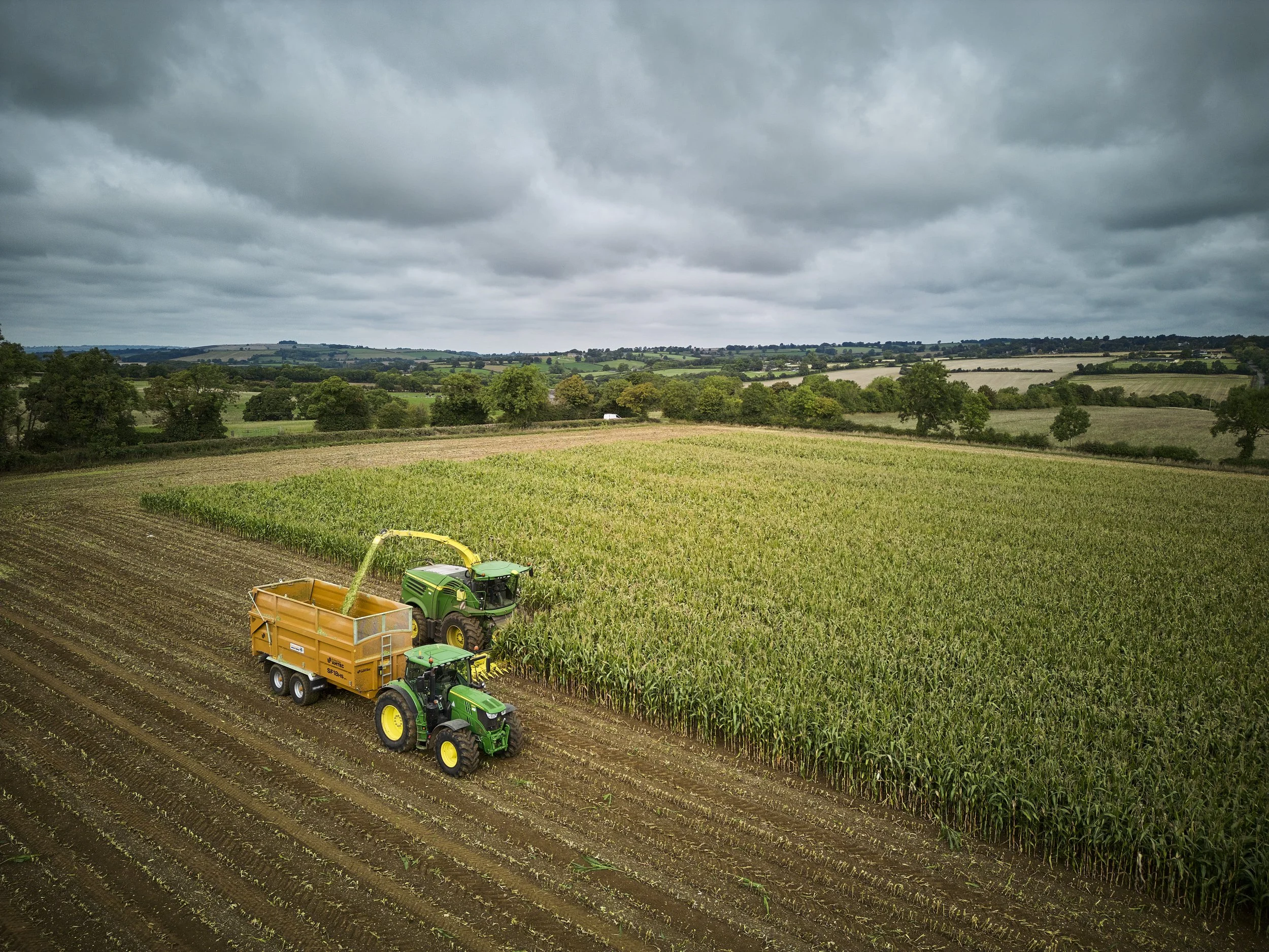 Tractors working on a cultivated farm field under cloudy sky with farm landscape and rolling hills in the background.
