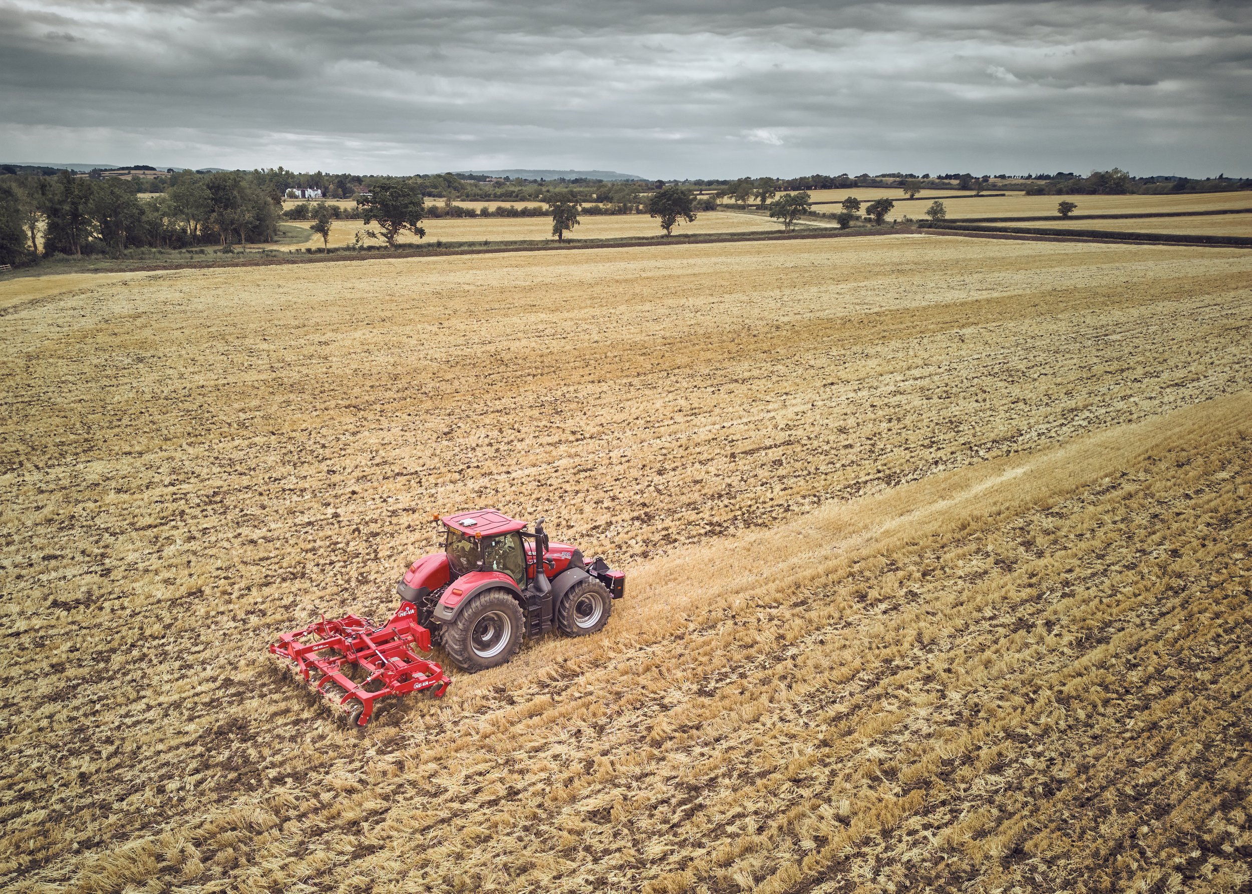 A red tractor working on a large, open field under a cloudy sky, with trees and distant fields visible in the background.