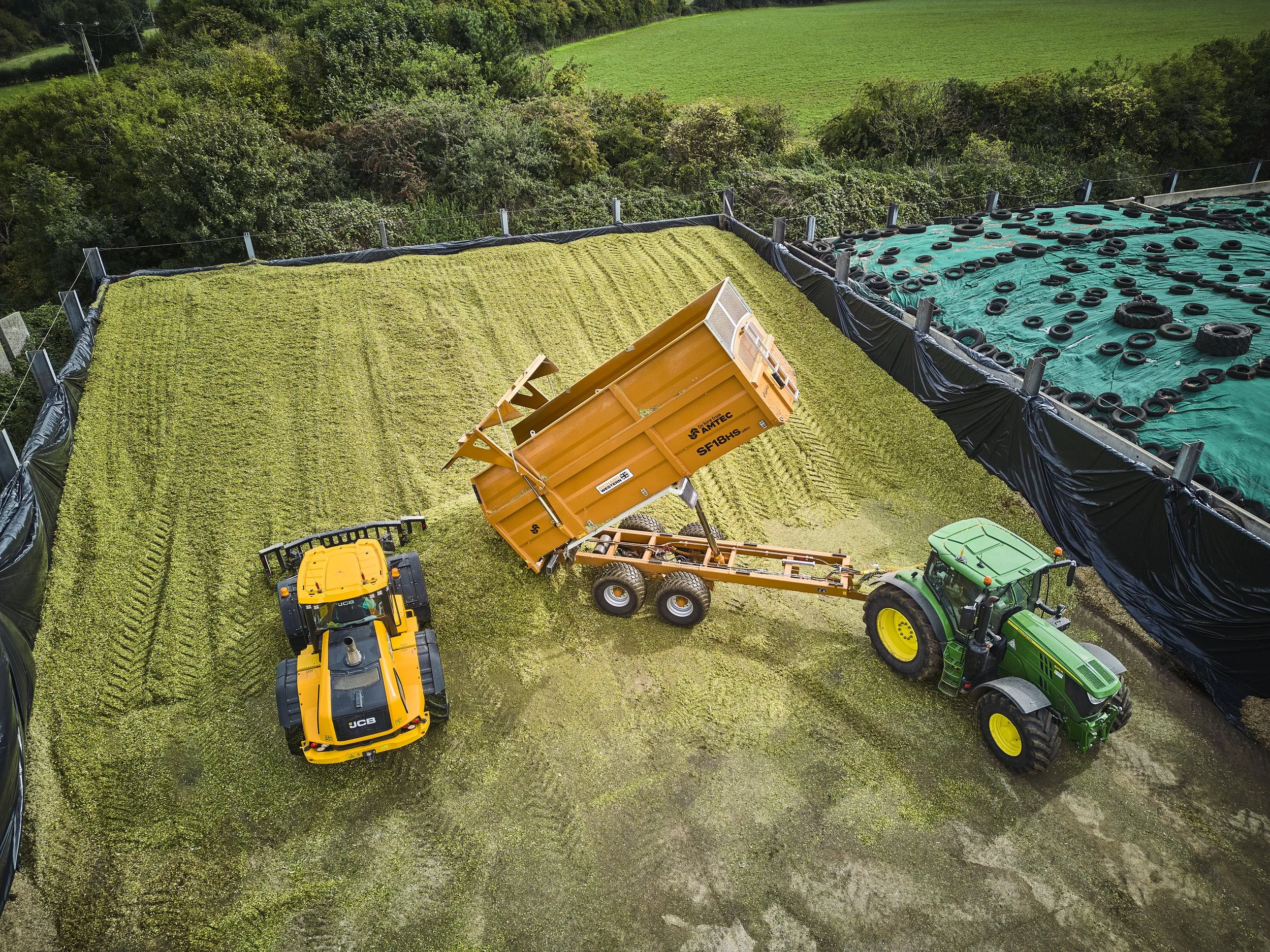 Aerial view of a green roof installation with construction equipment, including a yellow excavator, green tractor, and orange dump trailer, on a rooftop surrounded by trees and fields.