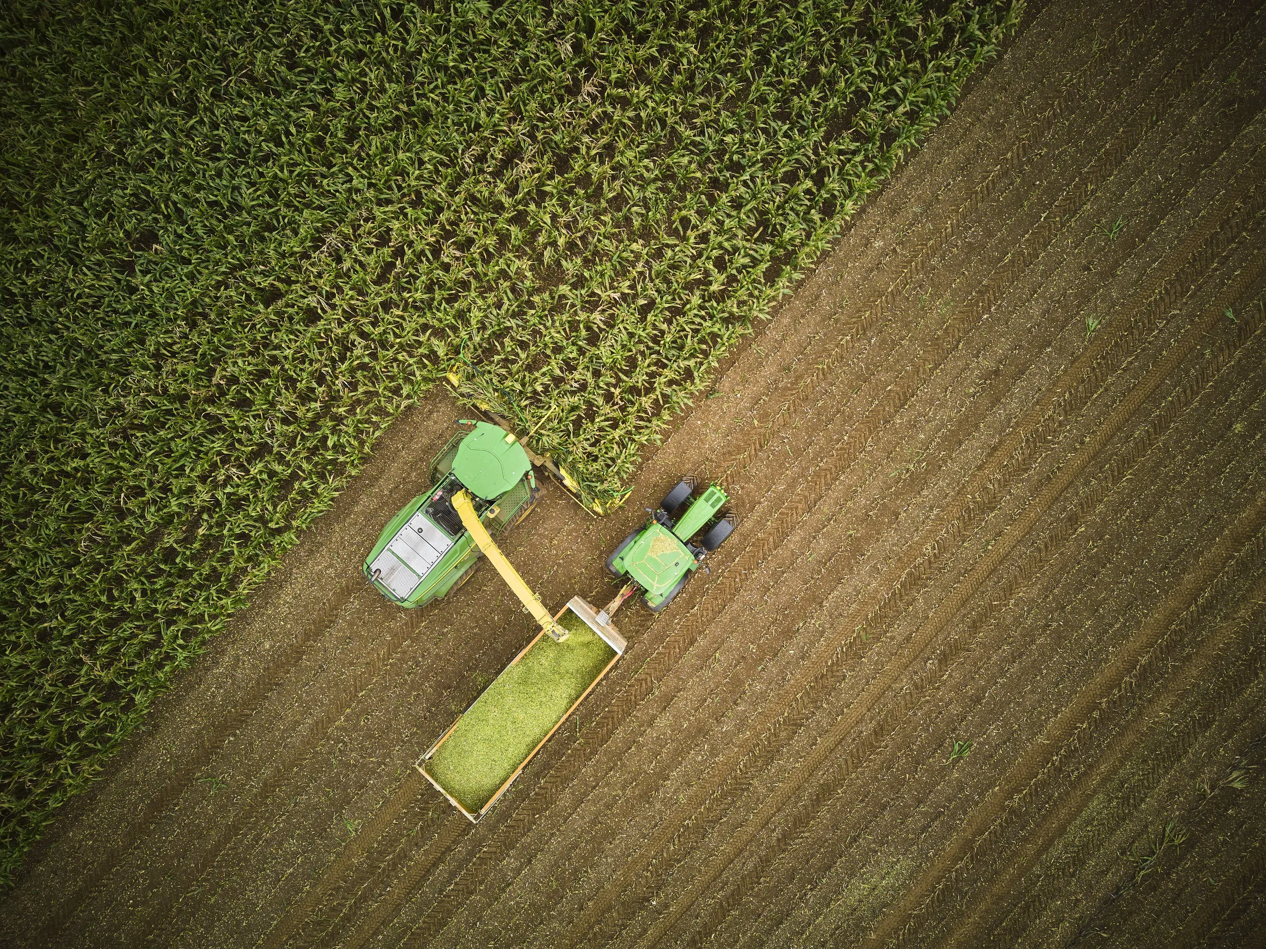 An aerial view of a farming field with green crops on the left and freshly tilled soil on the right. There is farming machinery, including a green tractor and equipment, working on the field.
