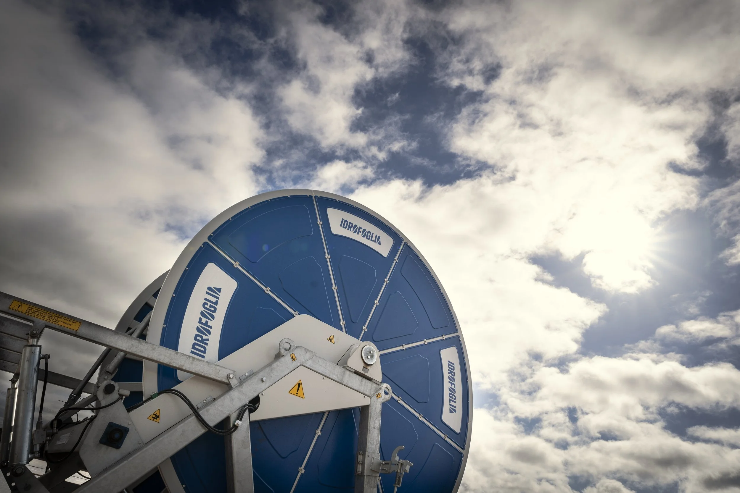 Large blue industrial cable reel labeled 'IDRAFGLIA' mounted on a metal structure, with a partly cloudy sky and sunlight in the background.