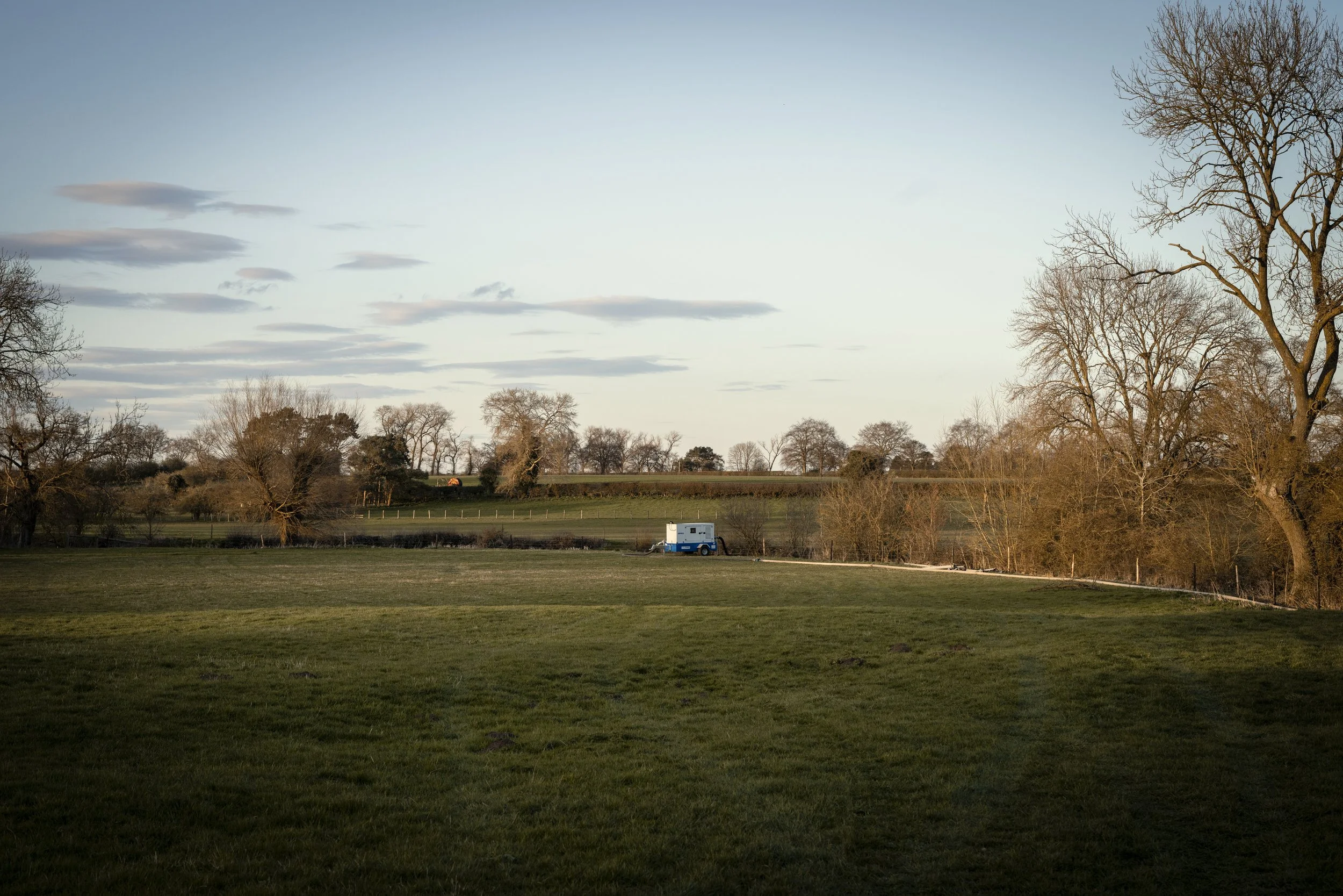 A peaceful rural landscape with a green grassy field, leafless trees, and a white and blue trailer in the distance during late afternoon or early evening.