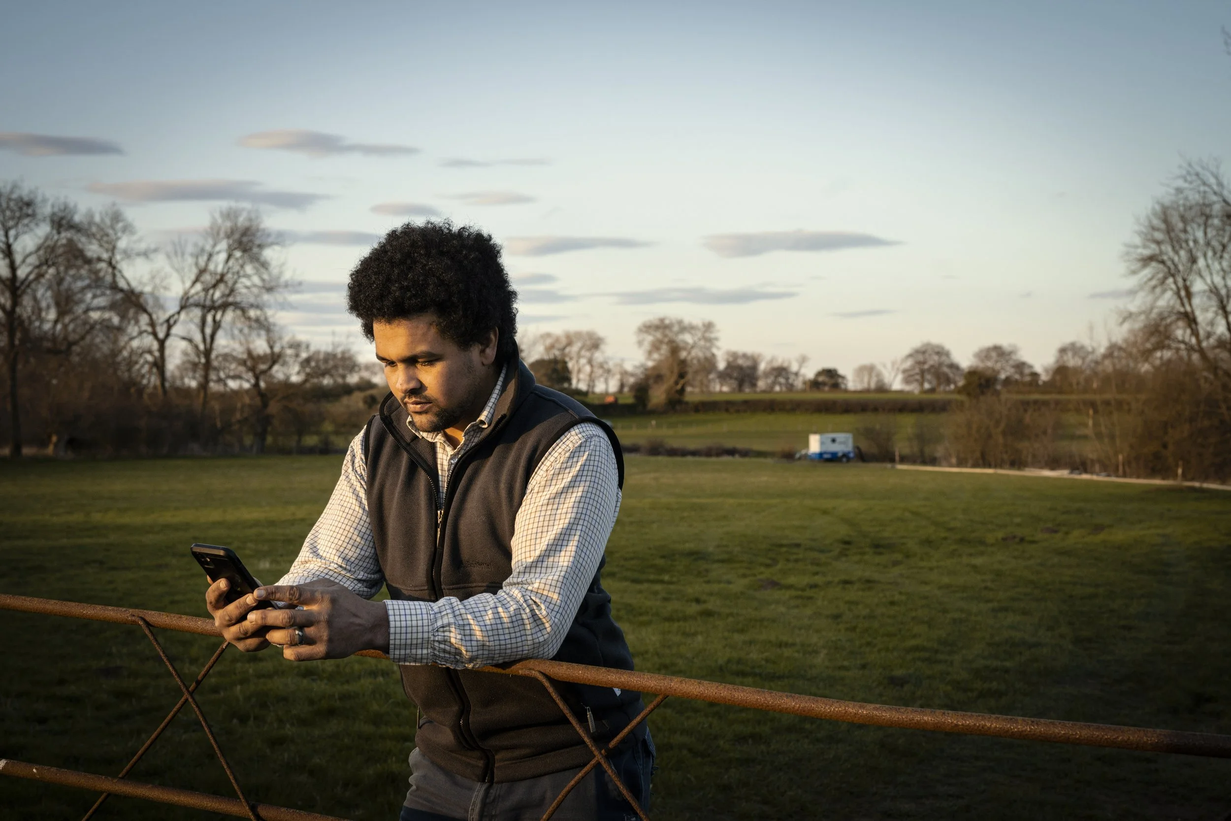 A man with curly black hair wearing a checkered shirt and black vest standing against a rusted metal fence in a rural field, looking at his smartphone as the sun sets in the background.
