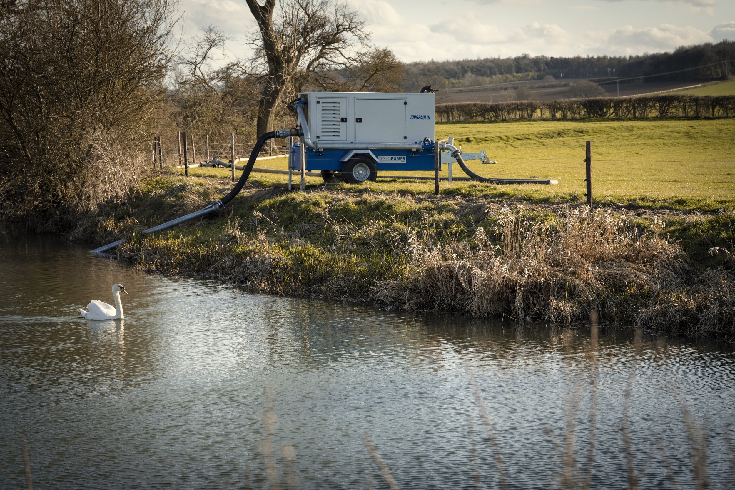 A water pump device positioned on a grassy riverbank, with two hoses extending into the water. A swan is swimming in the river in the foreground, and trees with no leaves are in the background on a sunny day.