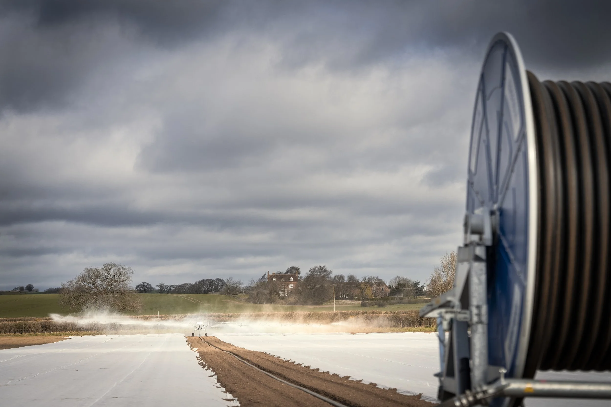 A large irrigation reel on the right side of the image with a farm field being watered under a cloudy sky.