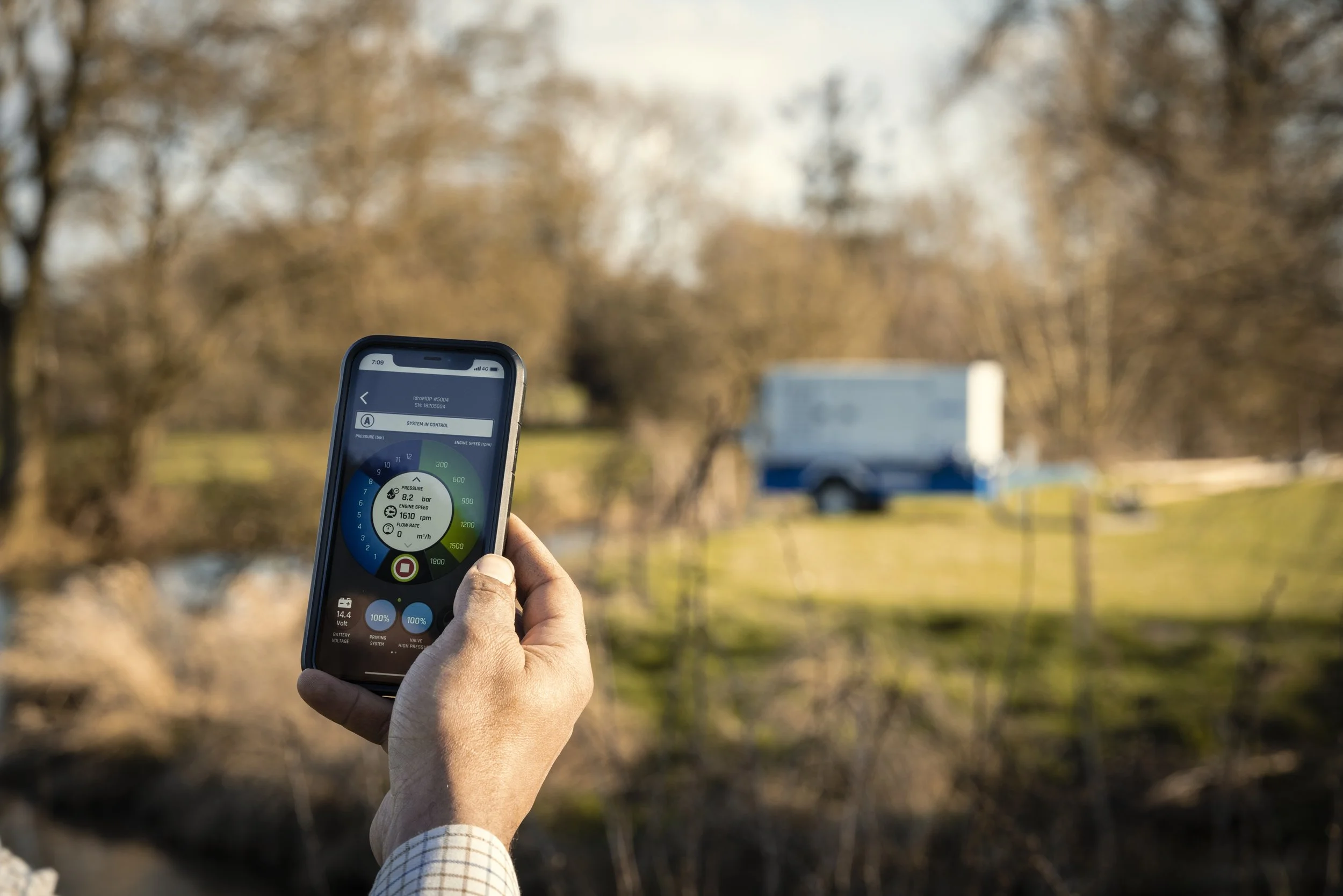 A person holding a smartphone with a vehicle monitoring app displayed, overlooking a blurred grassy area with trees and a blue and white truck in the background.