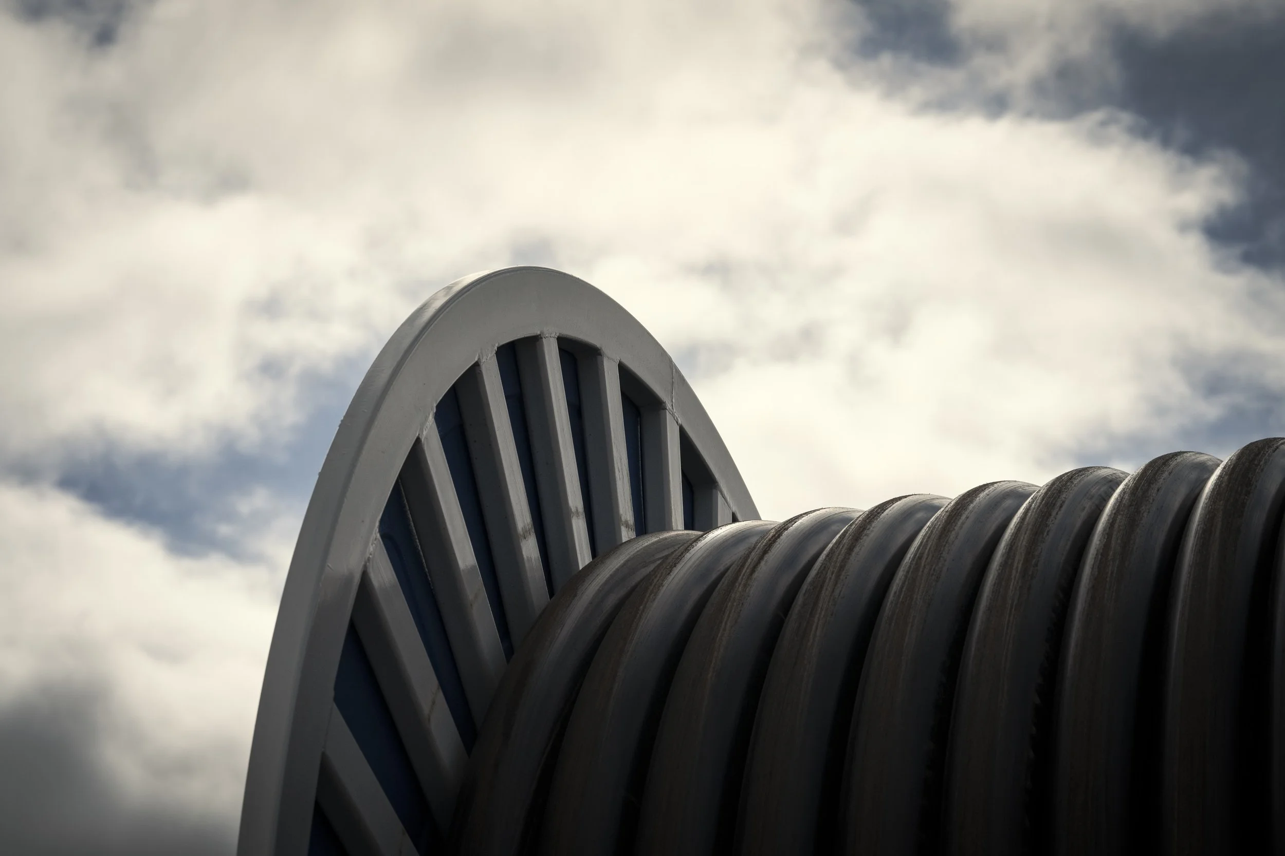 Close-up of a metallic, curved, slatted structure next to a large, curved, corrugated metal pipe against a cloudy sky.