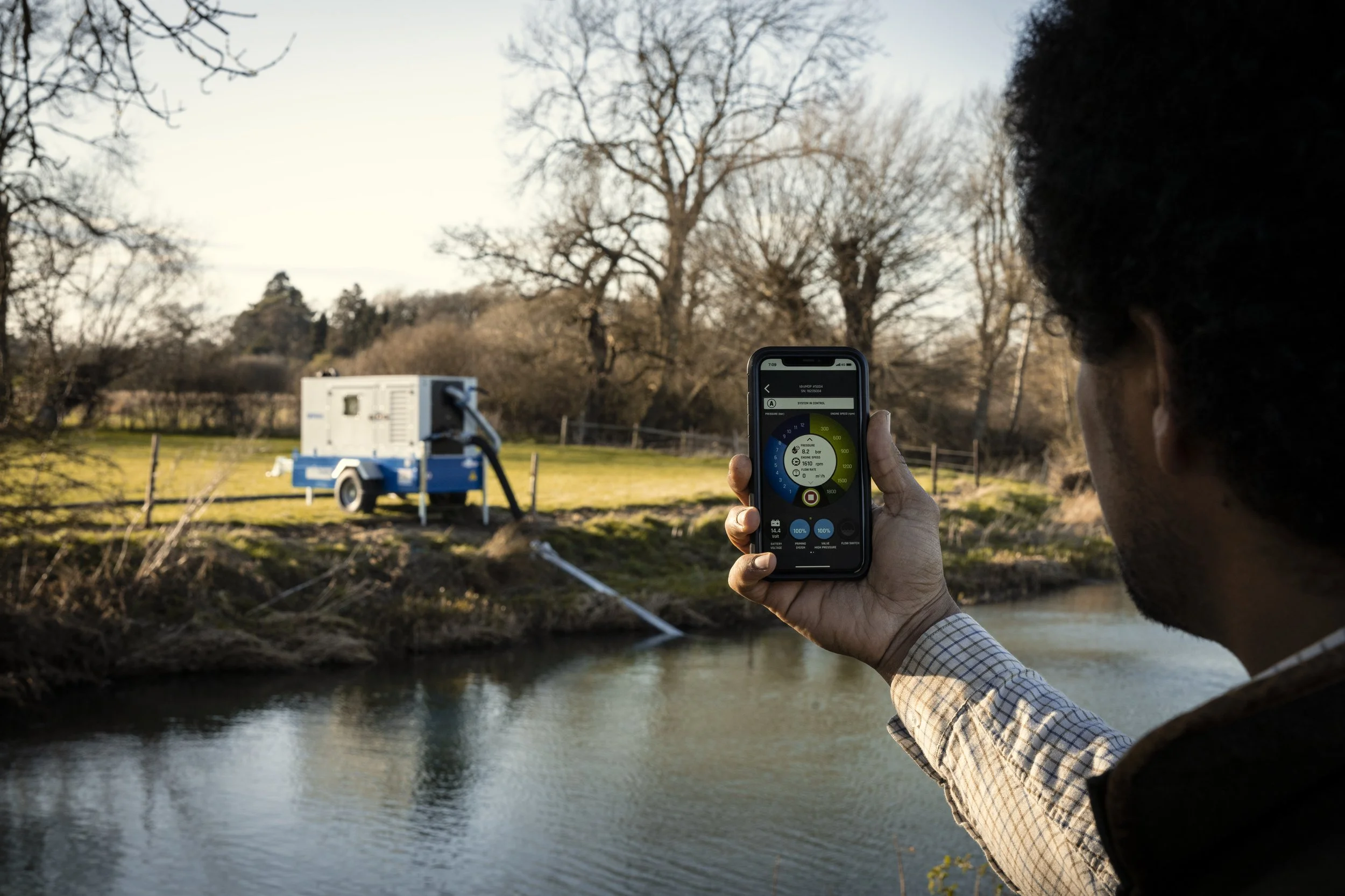 A person holding a smartphone with a control app open, facing a small blue and white generator near a river in a rural area during daylight.