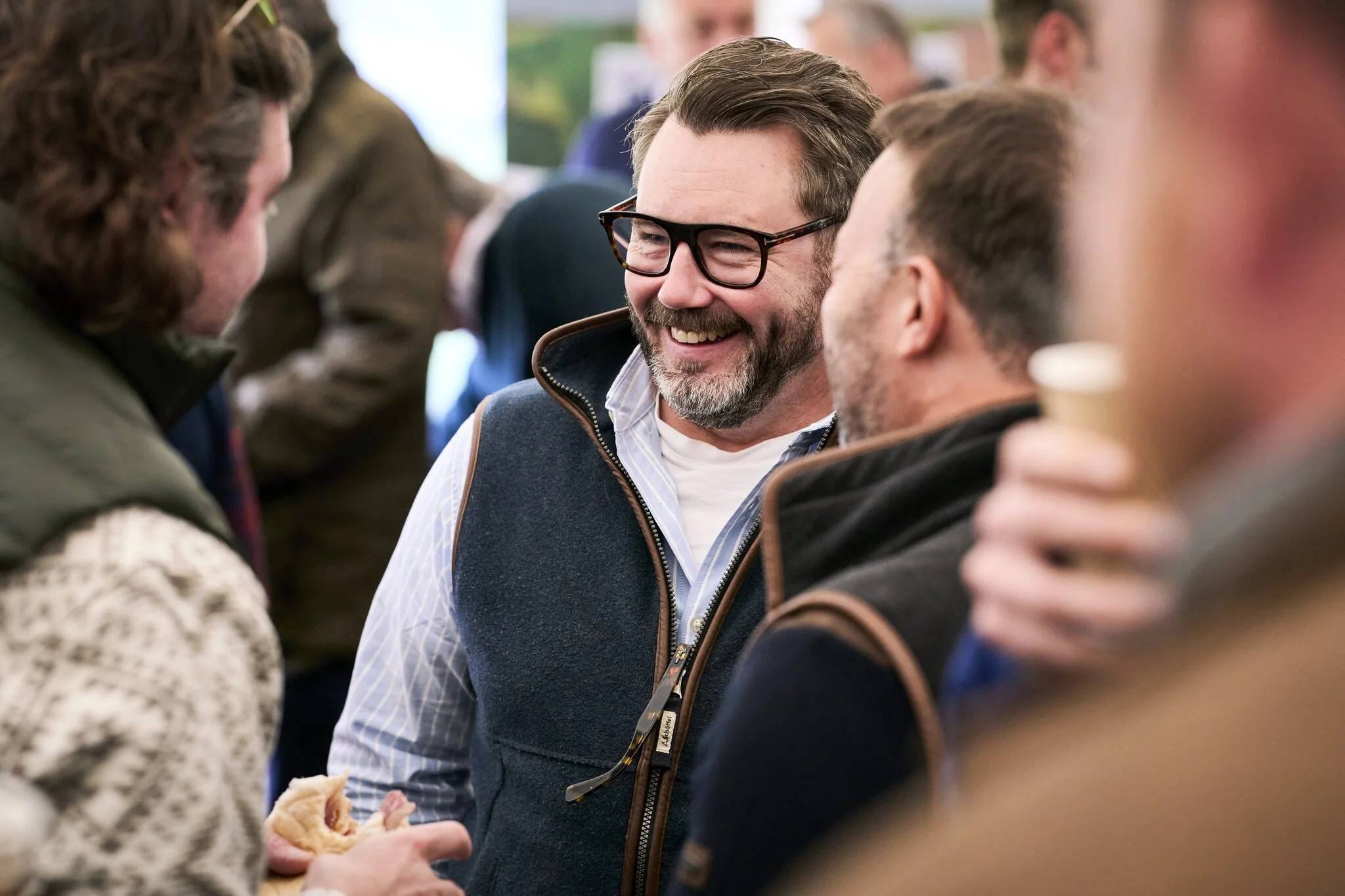 People gathered together in conversation, with one man smiling and wearing glasses at the center.