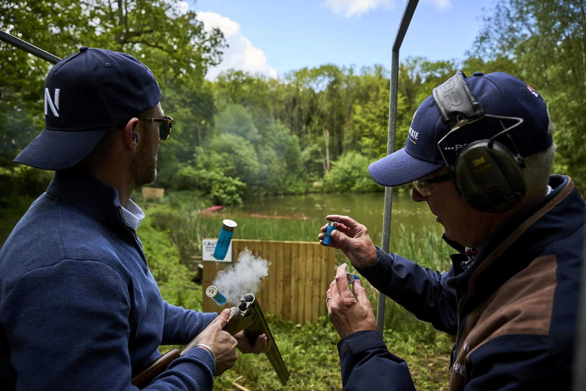 Two men preparing to set off fireworks outdoors near a pond, surrounded by greenery, with one man holding a firework and the other lighting it.