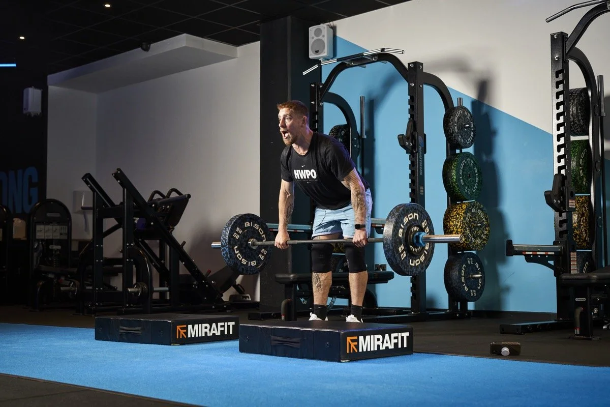 A man lifting a barbell during a workout in a gym. He is standing on a platform with the brand 'MIRAFIT'. The gym has weight plates and exercise equipment in the background.