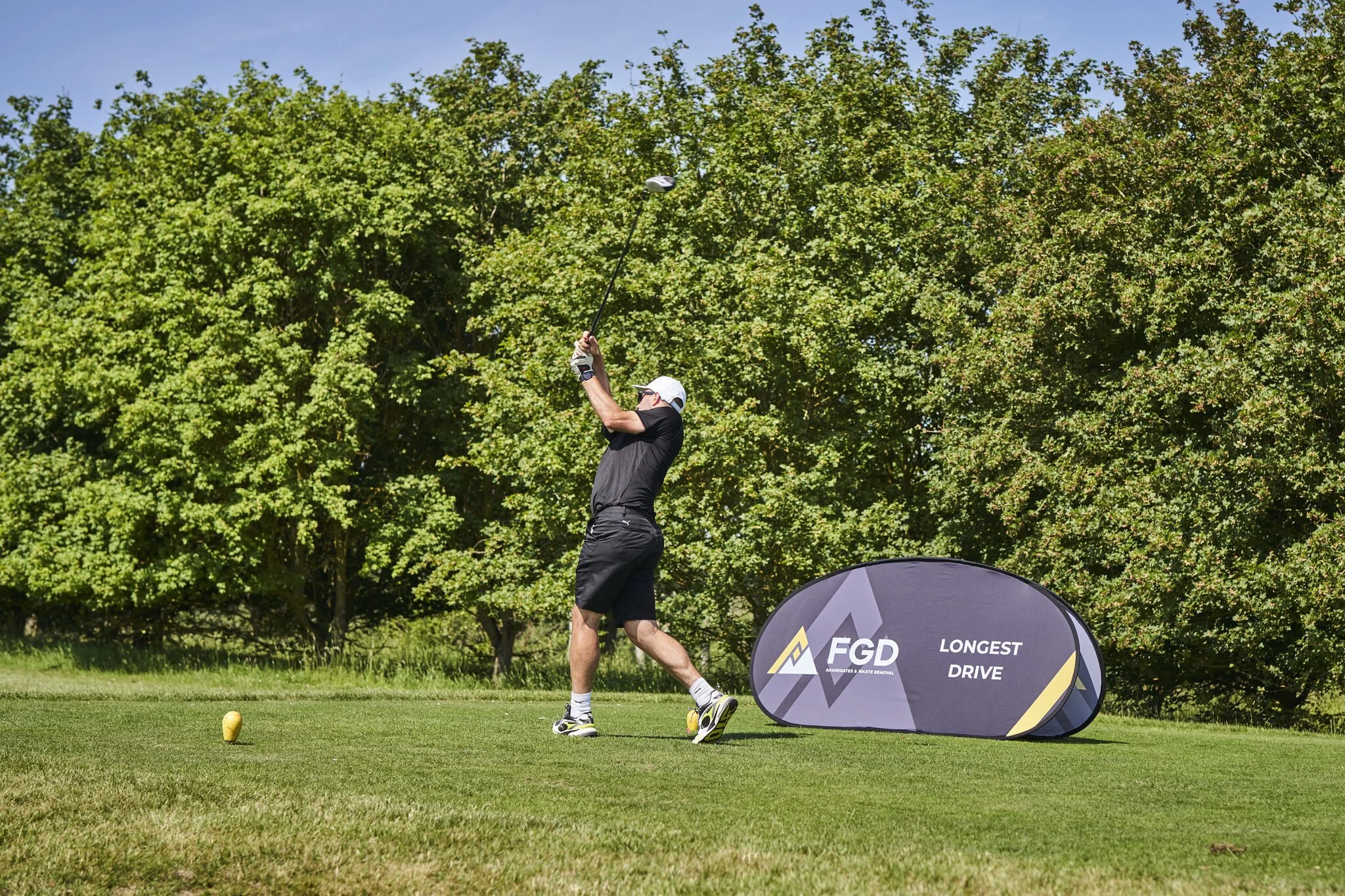 A man playing golf on a sunny day, mid-swing, with a golf club in his hands, standing on a golf course with green trees in the background, next to a black and yellow banner that reads 'FGD Longest Drive'.