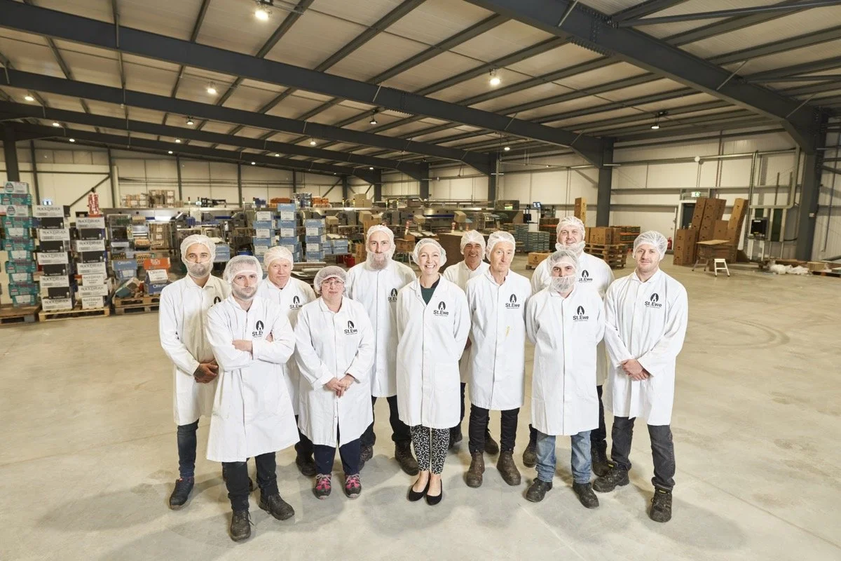Group of people in white lab coats and hairnets standing inside a large industrial warehouse with shelves stocked with boxes and supplies.