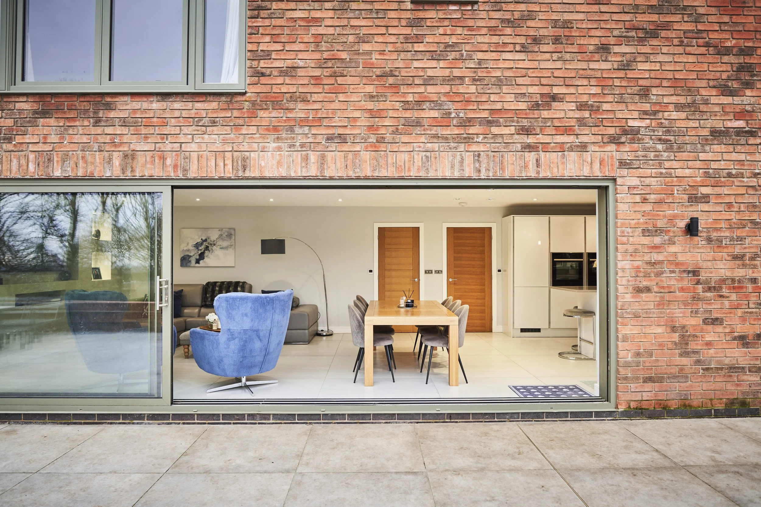 Interior view of a modern living room and dining area seen through a large sliding glass door. The room features a gray sofa, a blue armchair, a wooden dining table with six chairs, and contemporary kitchen appliances. The interior has white walls, m