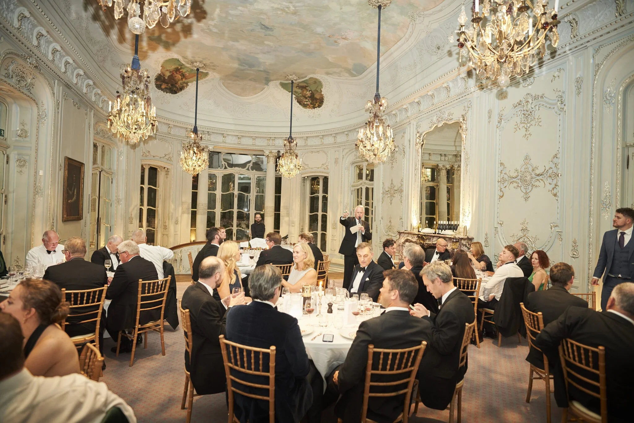 Elegant banquet hall with high ceilings, ornate chandeliers, and walls decorated with intricate moldings. Many guests in formal attire are seated at round tables, eating and conversing. A man in a suit stands and speaks to the audience.