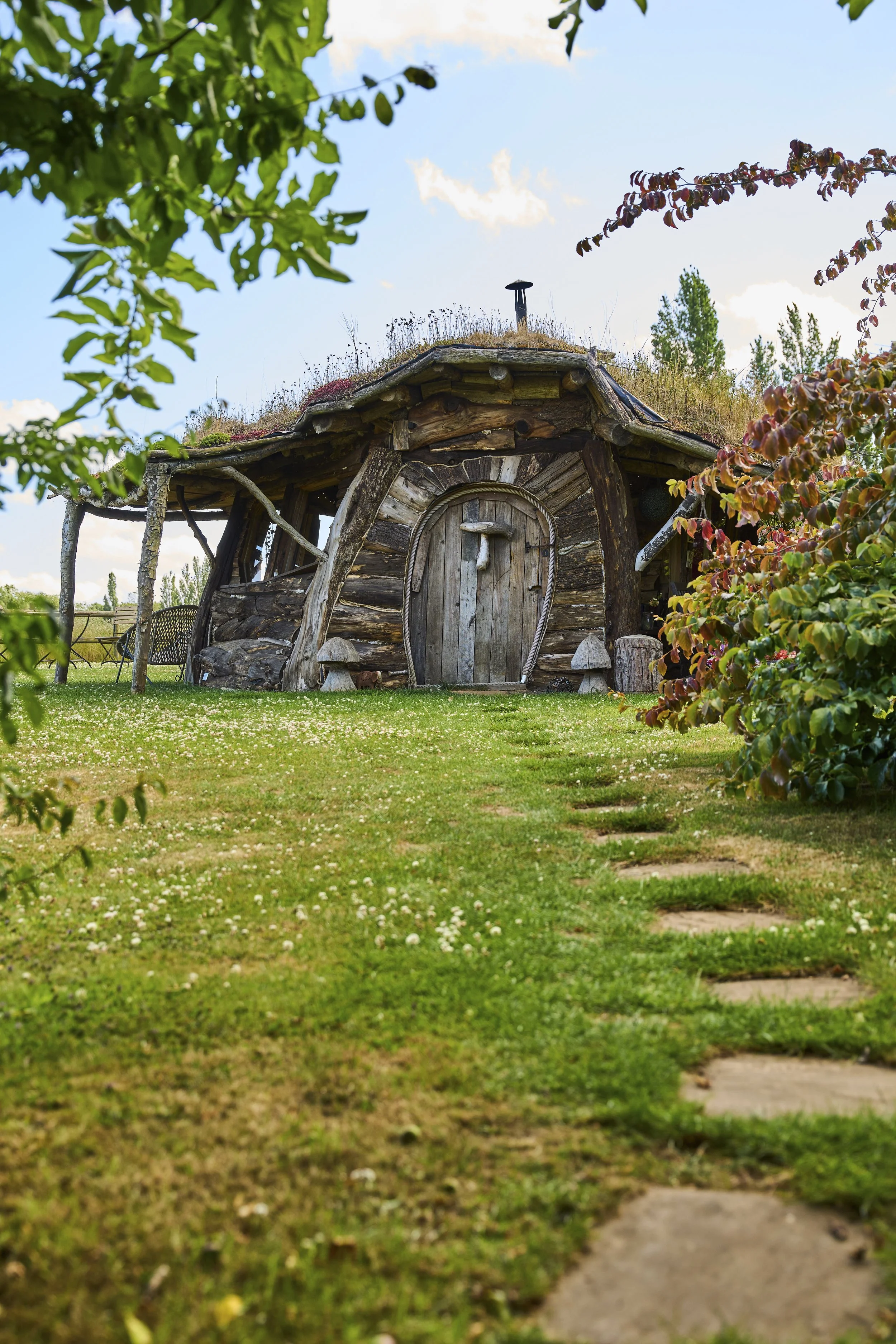 A rustic wooden hobbit house with a grass roof, surrounded by trees and a green lawn, with a stepping stone pathway leading to the door.