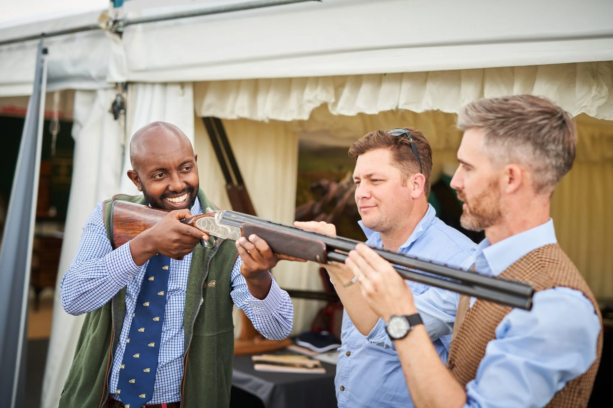 Three men are outdoors in front of a tent, with one man aiming and firing a shotgun while the other two hold the shotgun steady.