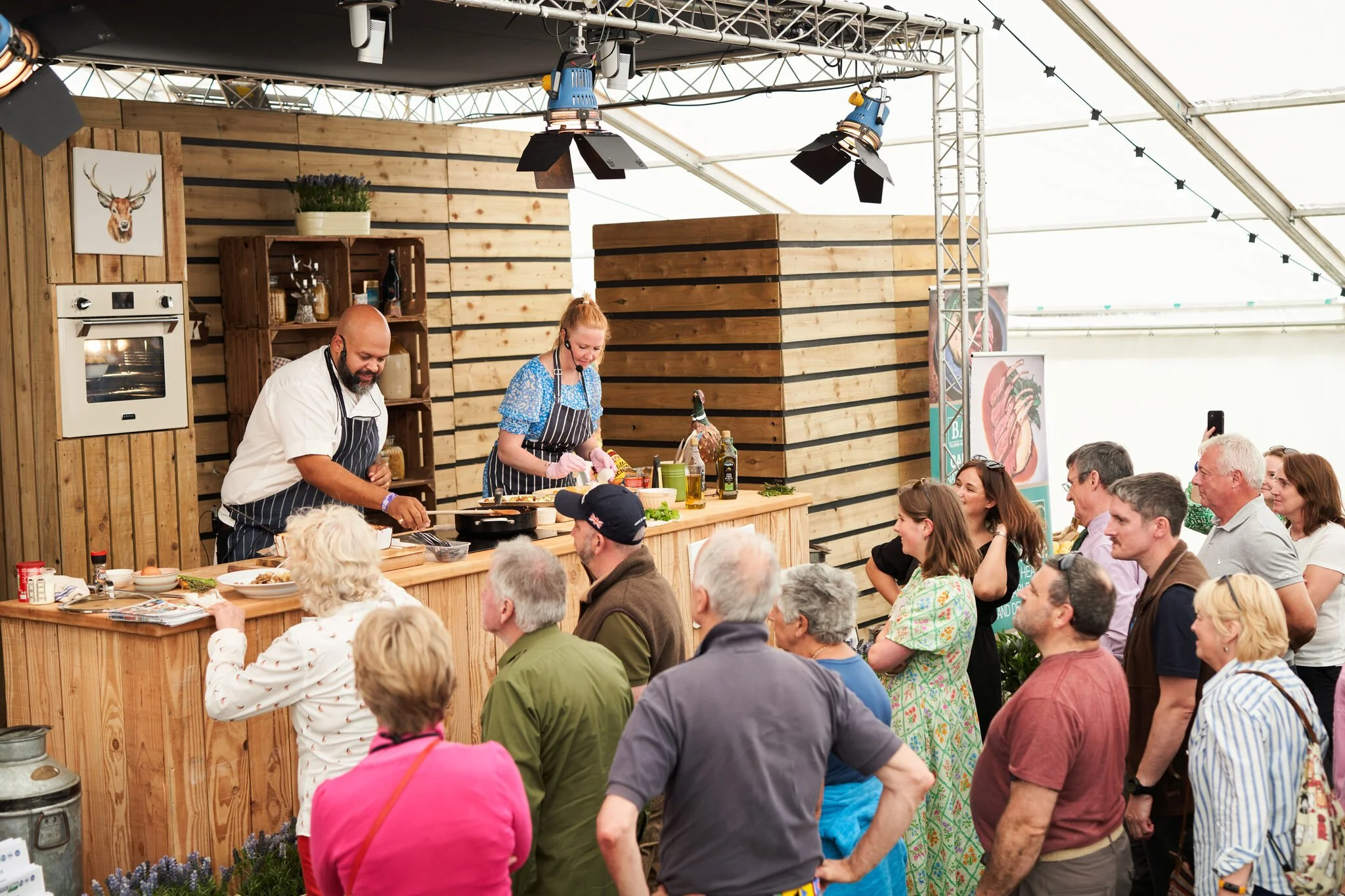 People gathered at a cooking demonstration in a large tent with wooden walls and stage lighting. Two chefs are preparing food at a wooden counter, surrounded by an audience.