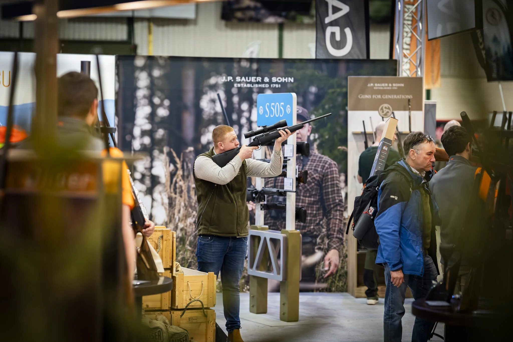 A man with a red hair and beard holding a scoped rifle in a shooting stance at an indoor gun exhibition. Other attendees and display booths are visible in the background.