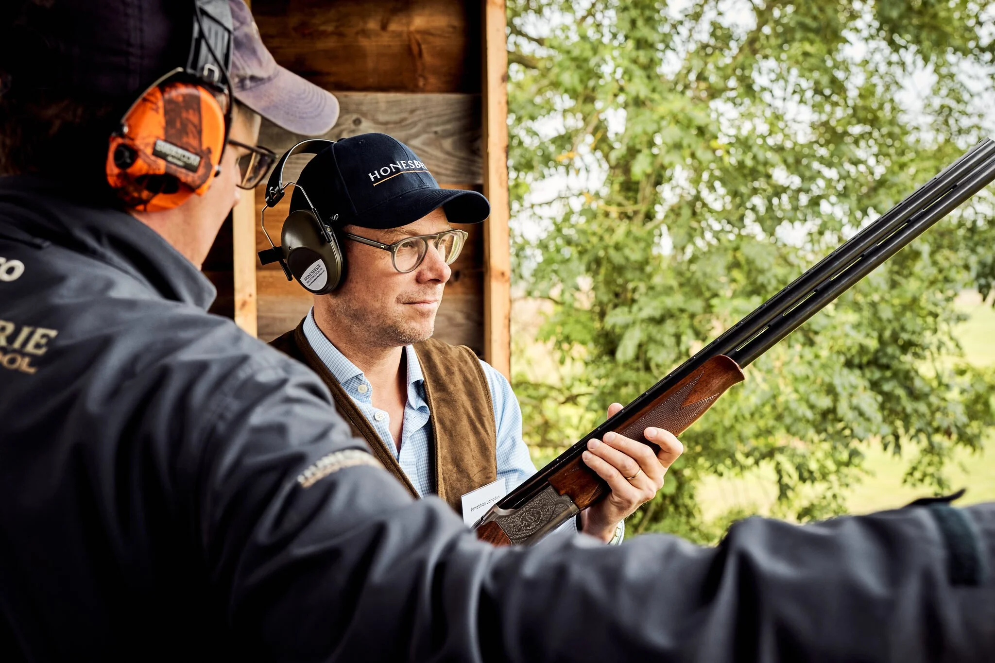 Two men wearing ear protection and hats, one with glasses, outdoors, with trees in the background. One man is holding a rifle, and they appear to be engaged in shooting or firearm training.