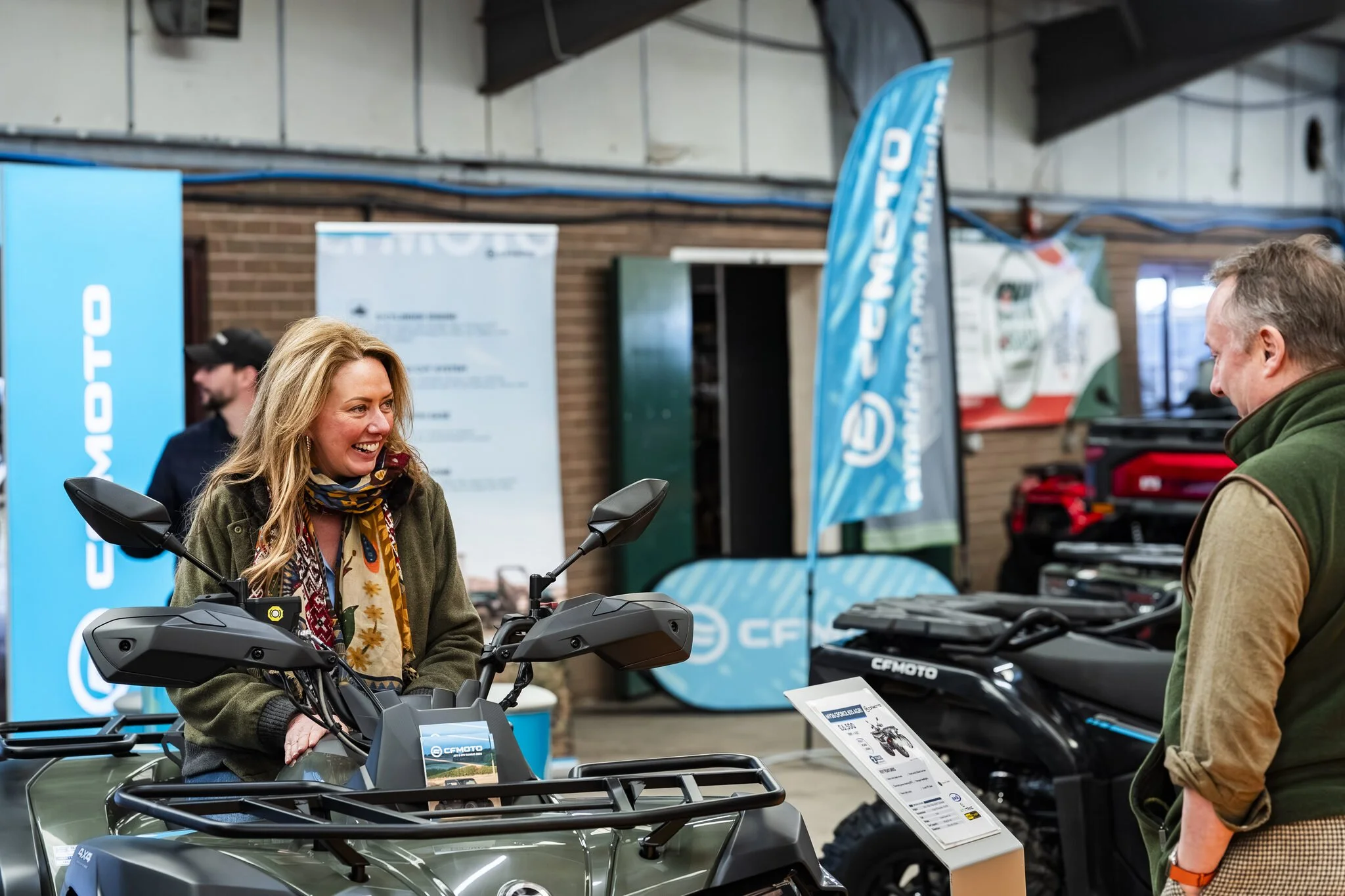 A woman with blonde hair smiling while sitting on an ATV at an indoor showroom, talking to a man. Blue banners and information stands about ATVs and outdoor equipment are visible in the background.
