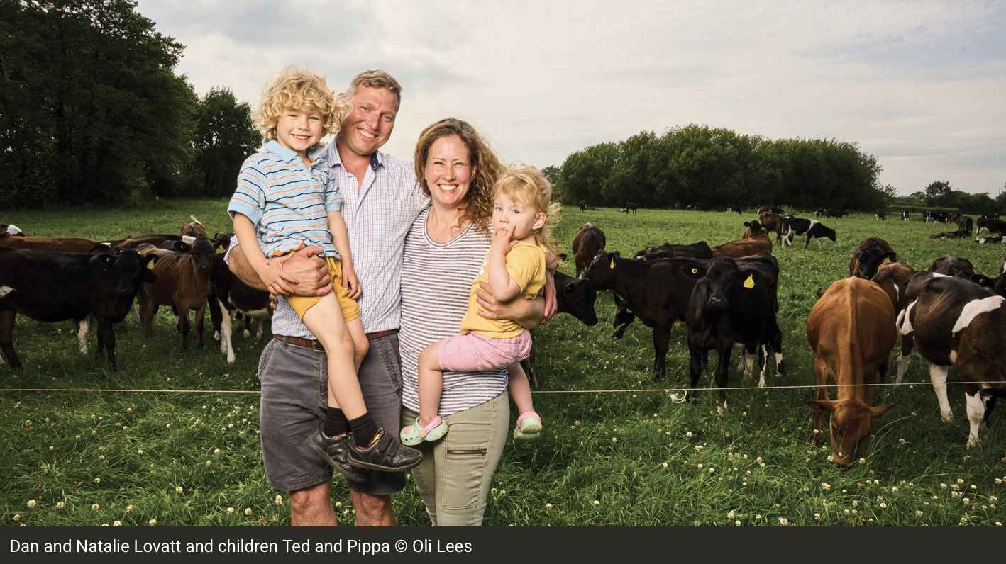 Family of four standing in a field with cows, smiling, with trees and a cloudy sky in the background.