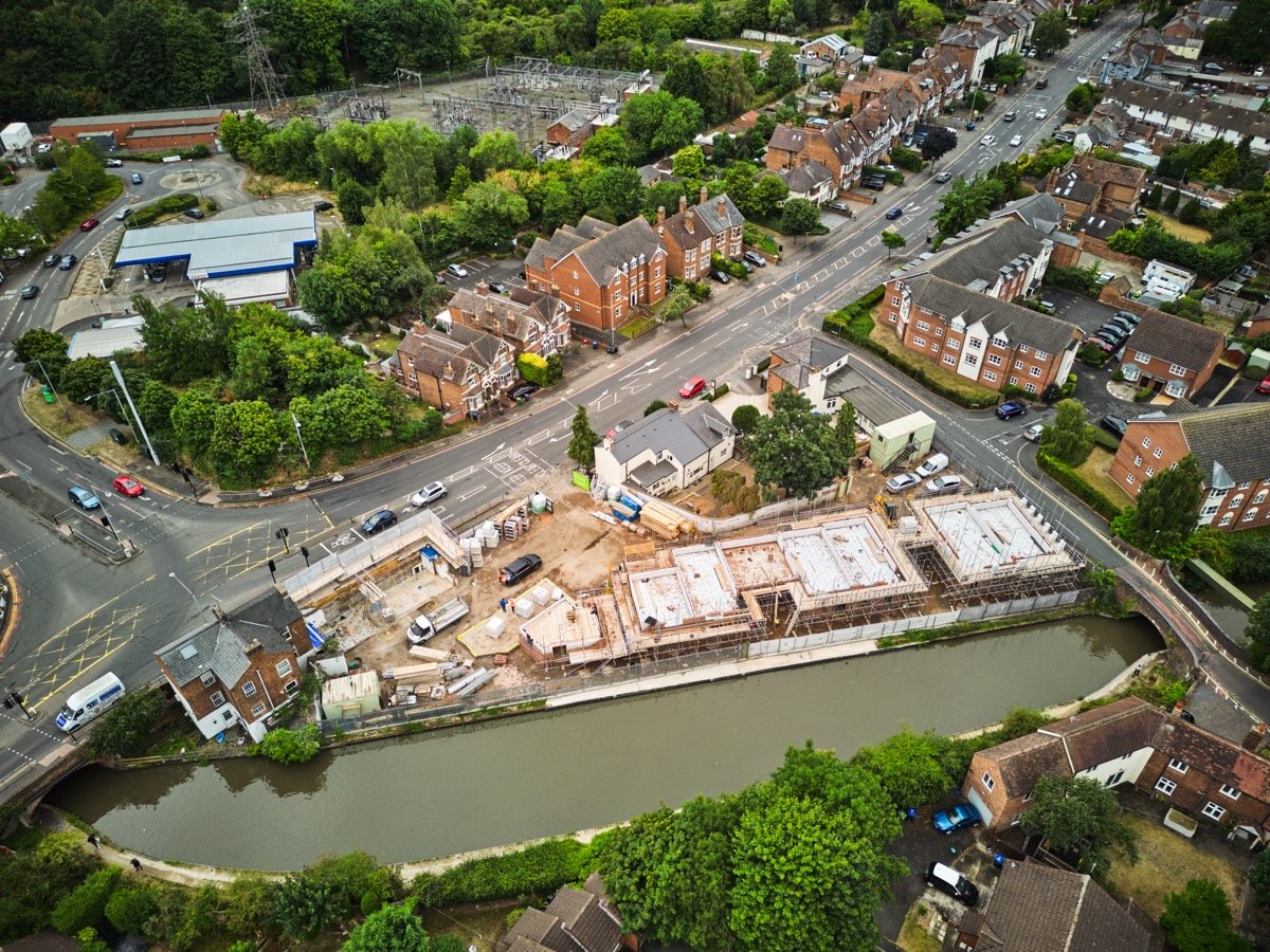 Aerial view of a construction site near a river, surrounded by residential houses and a wooded area, with roads and parked cars visible.