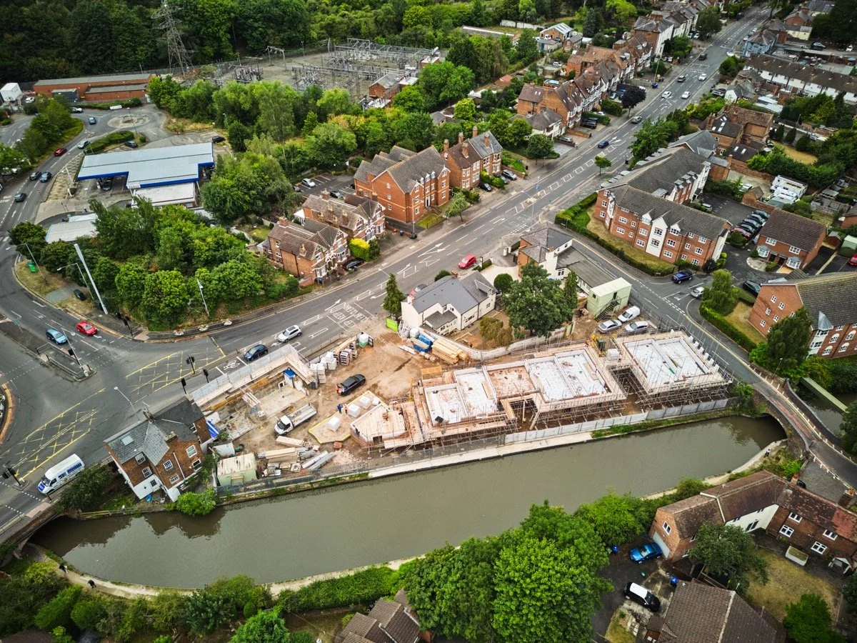 Aerial view of a construction site near a river, surrounded by residential houses and a wooded area, with roads and parked cars visible.