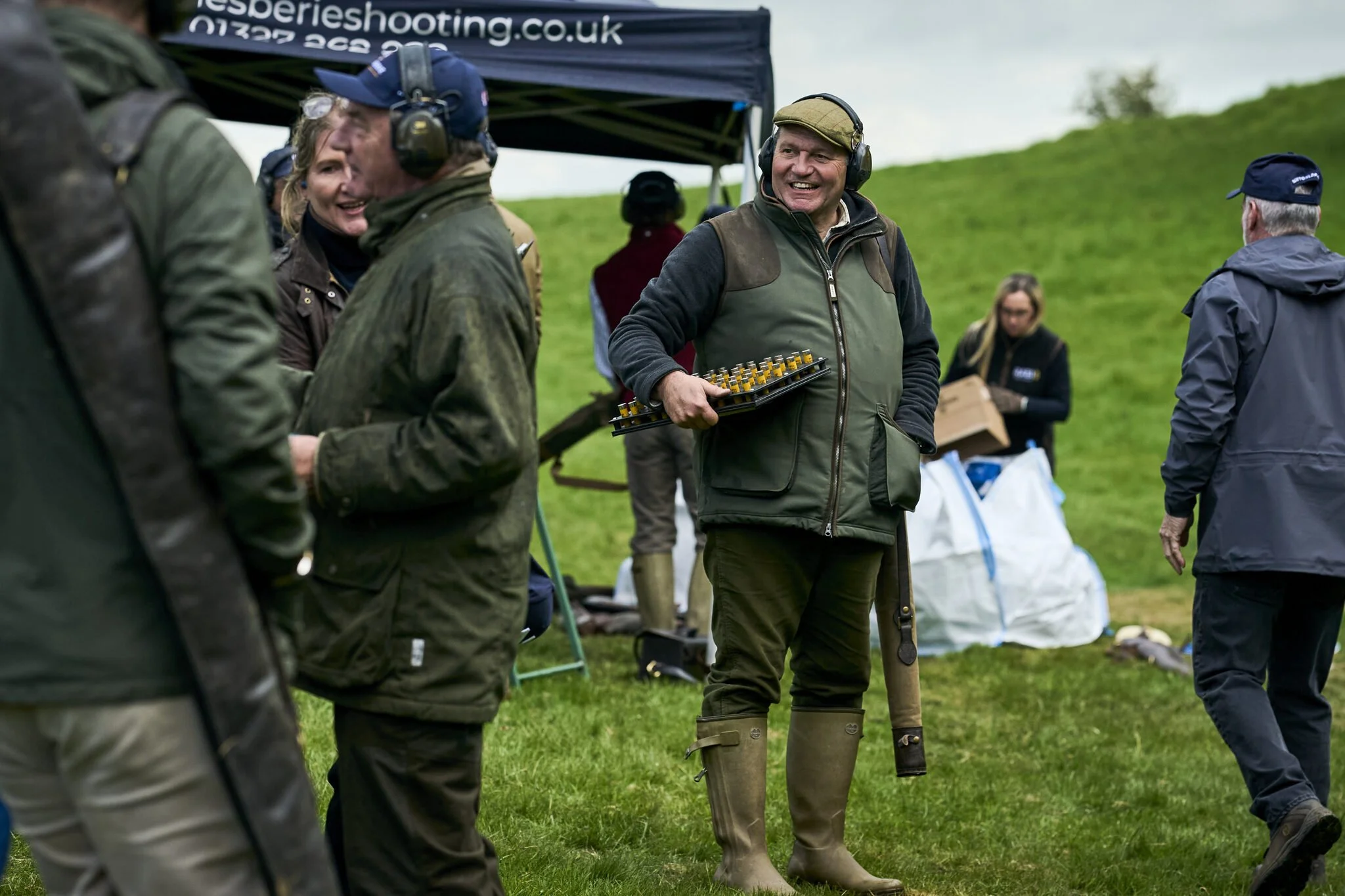 People outdoors at an event, some smiling, wearing headphones, jackets, and boots, with a tent in the background.
