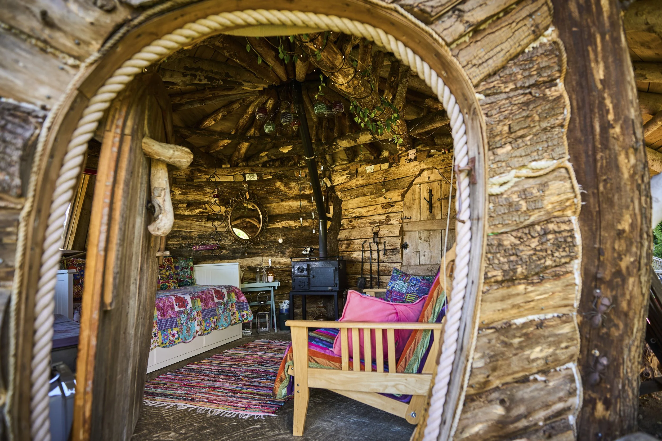 Interior of a rustic wooden log cabin with cozy furnishings, including a wood stove, colorful patchwork bedspread, and a pink cushion armchair, seen through an arched doorway with rope trim.