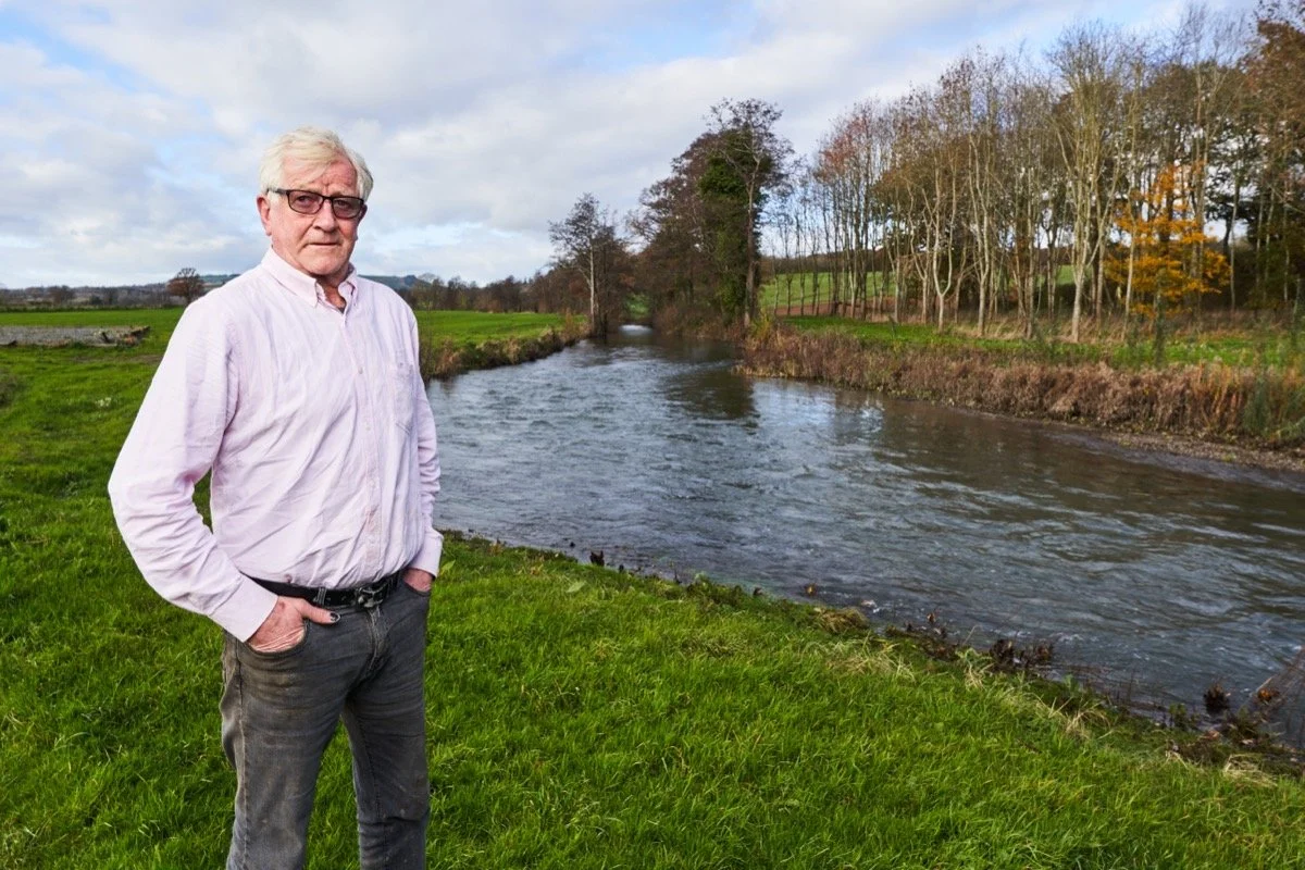 An older man wearing glasses and a light pink button-up shirt stands on green grass near a river with trees in the background, under a partly cloudy sky.