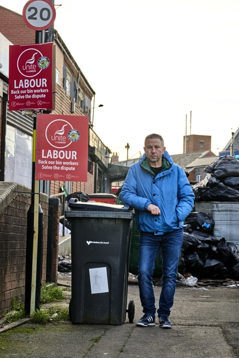 A man in a blue jacket standing next to a black trash bin on a city sidewalk, with signs supporting labor rights and a pile of black garbage bags behind him.