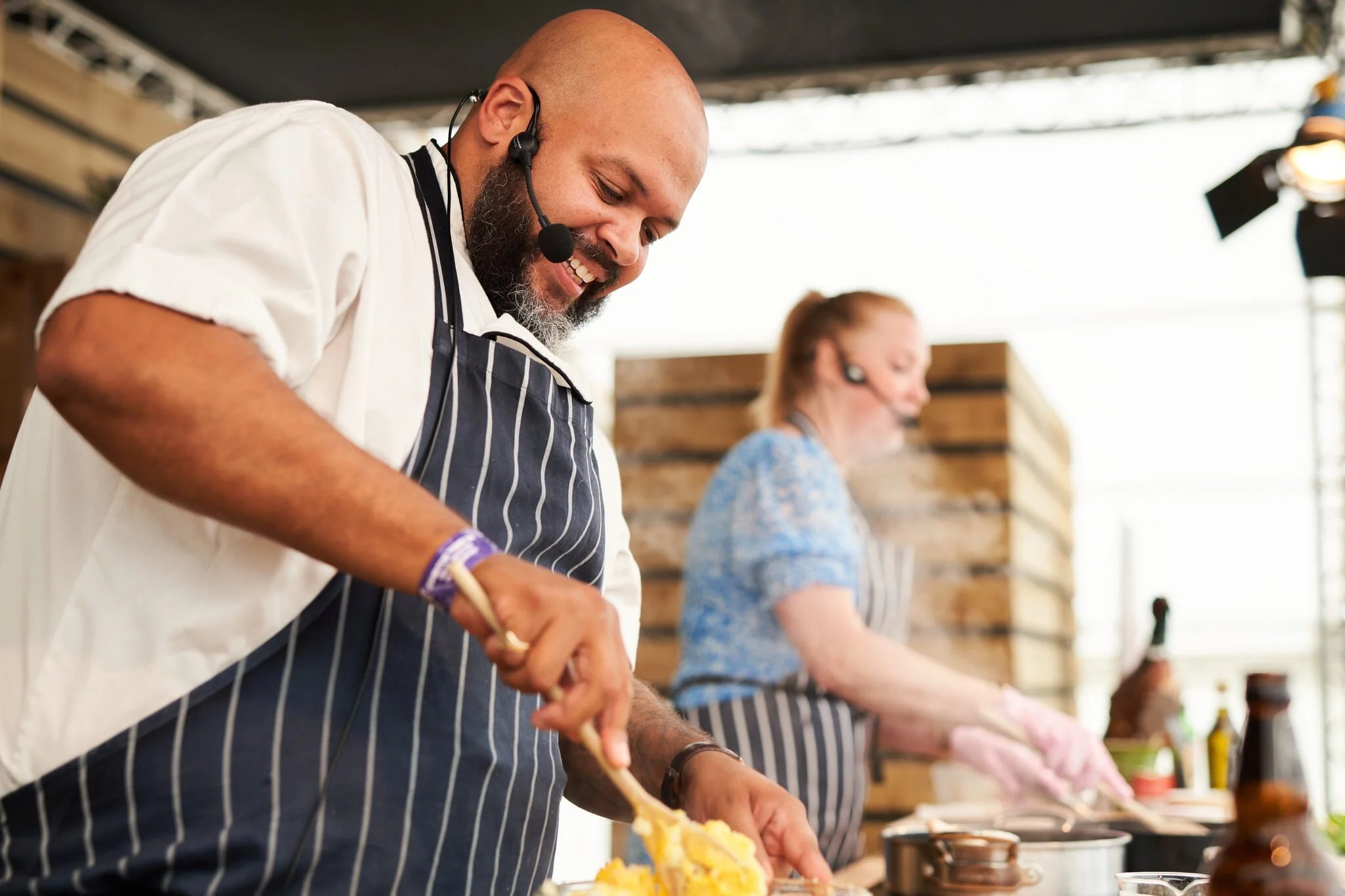 A man with a bald head and beard wearing a headset and an apron is cooking or preparing food at a counter with multiple people. The person beside him, a woman with light-colored hair, is also working on food. The background includes a wooden wall and