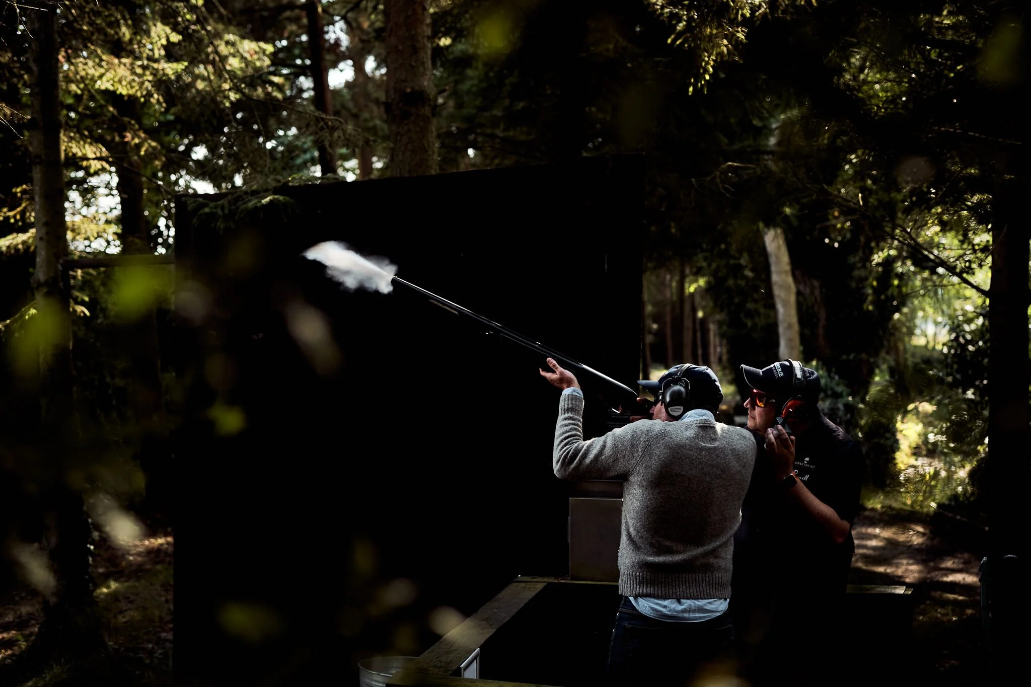 People in a forest setting appear to be involving in some activity with a device emitting smoke or vapor, possibly a smoke machine or similar equipment, with trees and natural light visible in the background.
