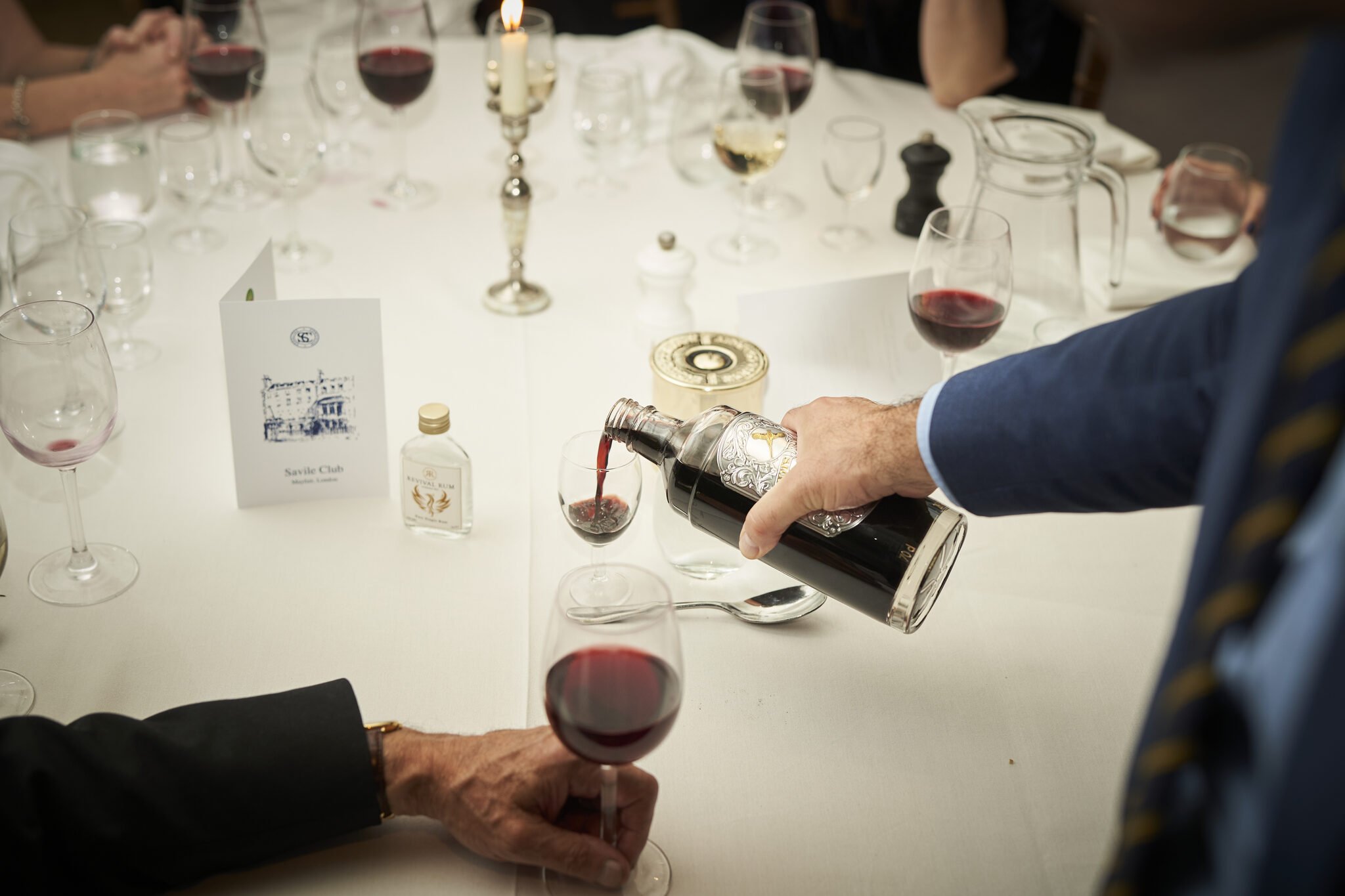 A person in a suit pouring red wine into a glass at a formal dinner table, surrounded by various glasses, candles, and bottles.