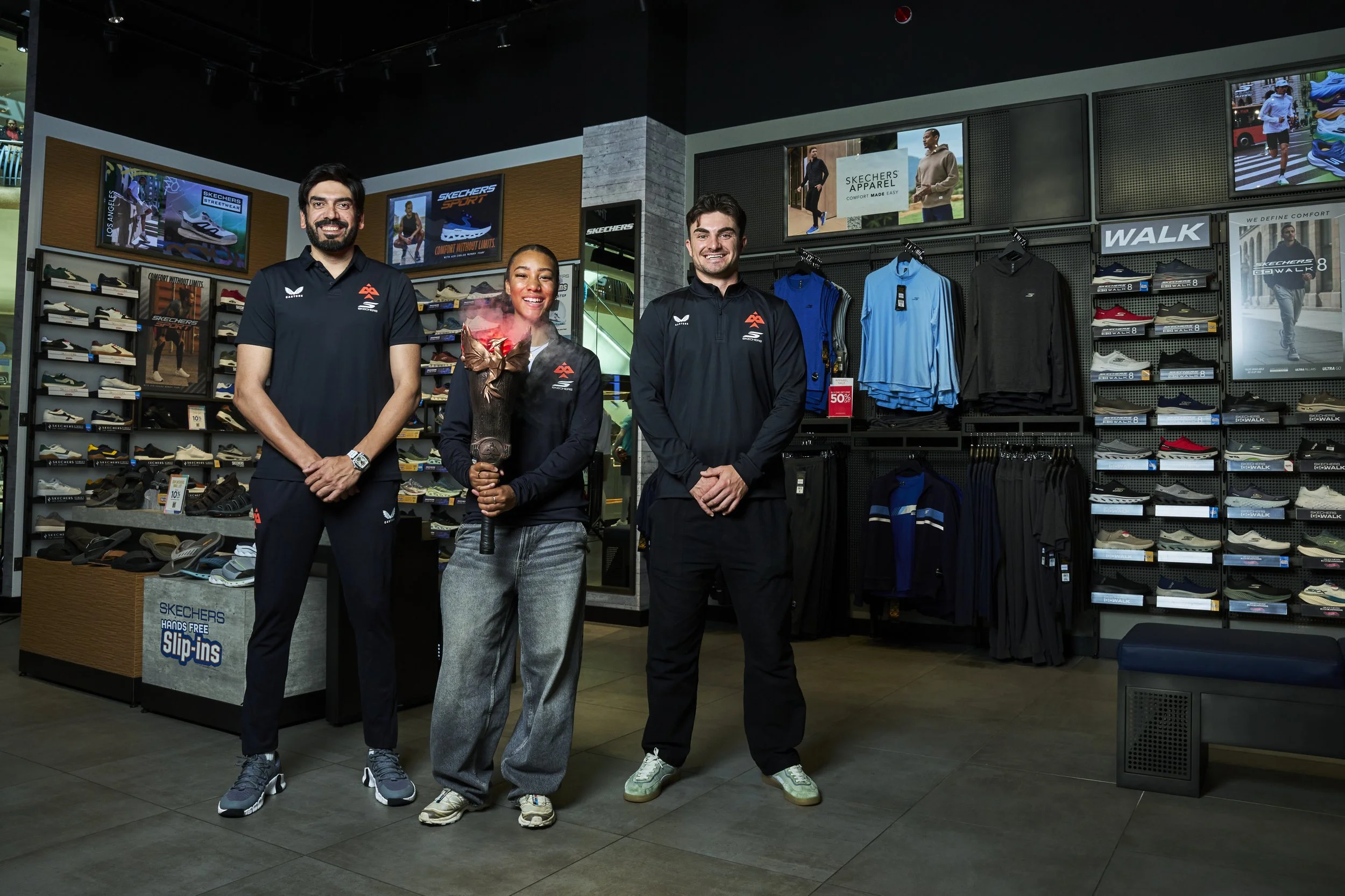 Three people standing inside a shoe store, smiling at the camera. Shelves behind them display various athletic shoes and apparel, with digital advertisements and posters on the walls.