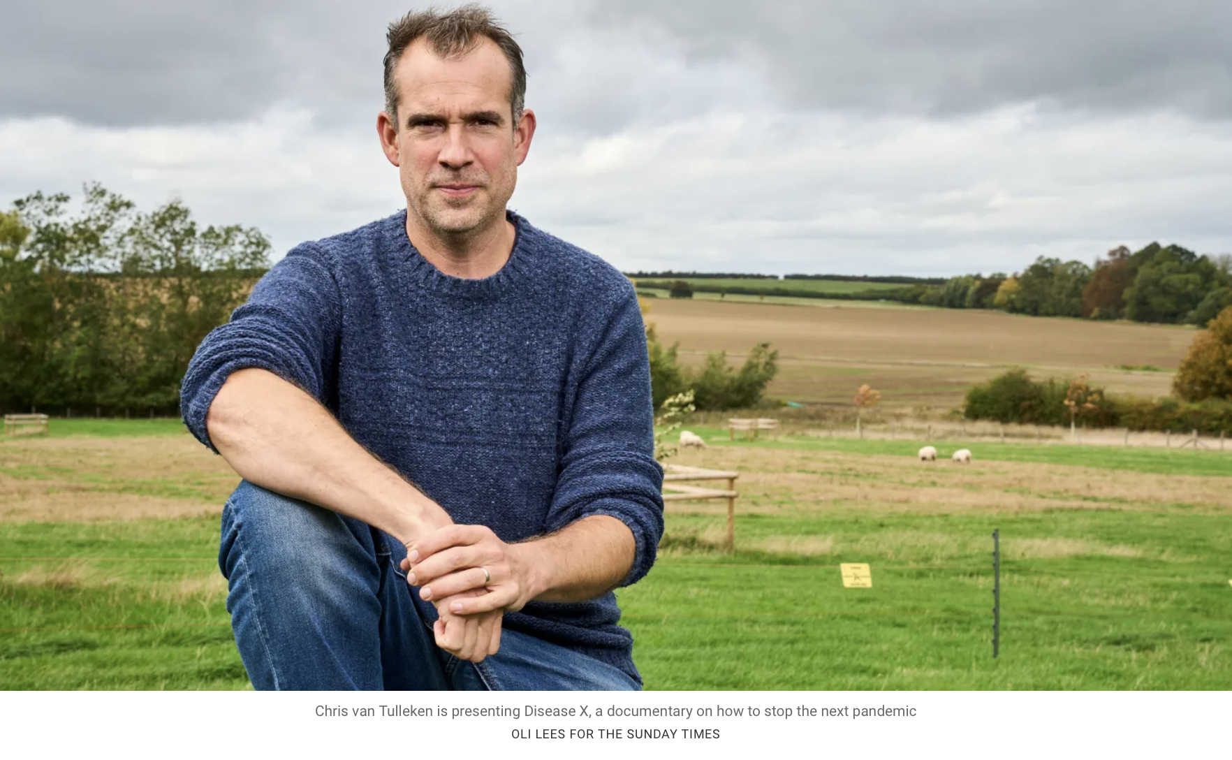 Chris van Tulleken kneeling outdoors on a grassy field with rolling hills and sheep in the background.