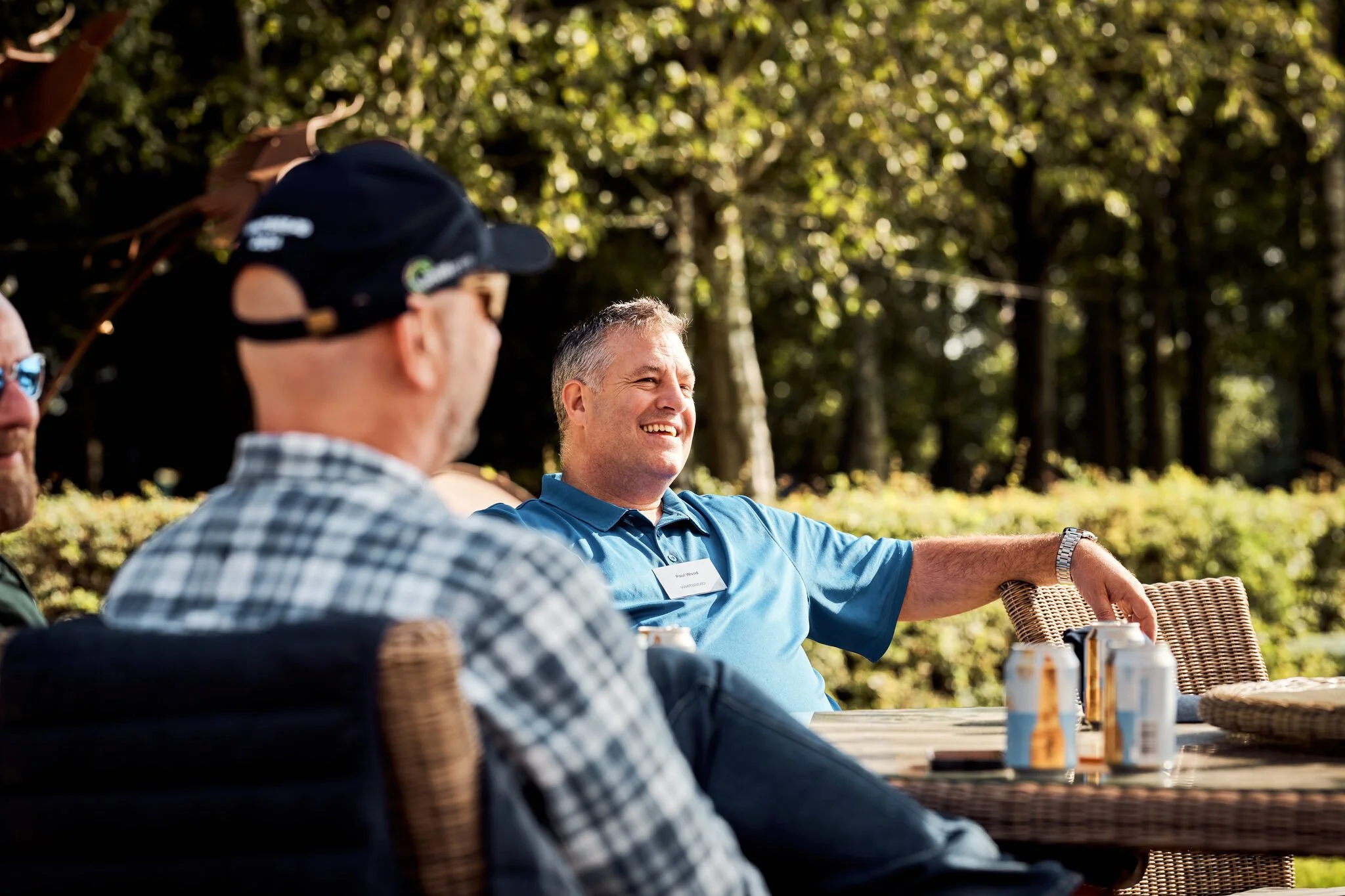 Group of men sitting outdoors at a table, enjoying a sunny day, with some drinks and snacks, surrounded by trees.